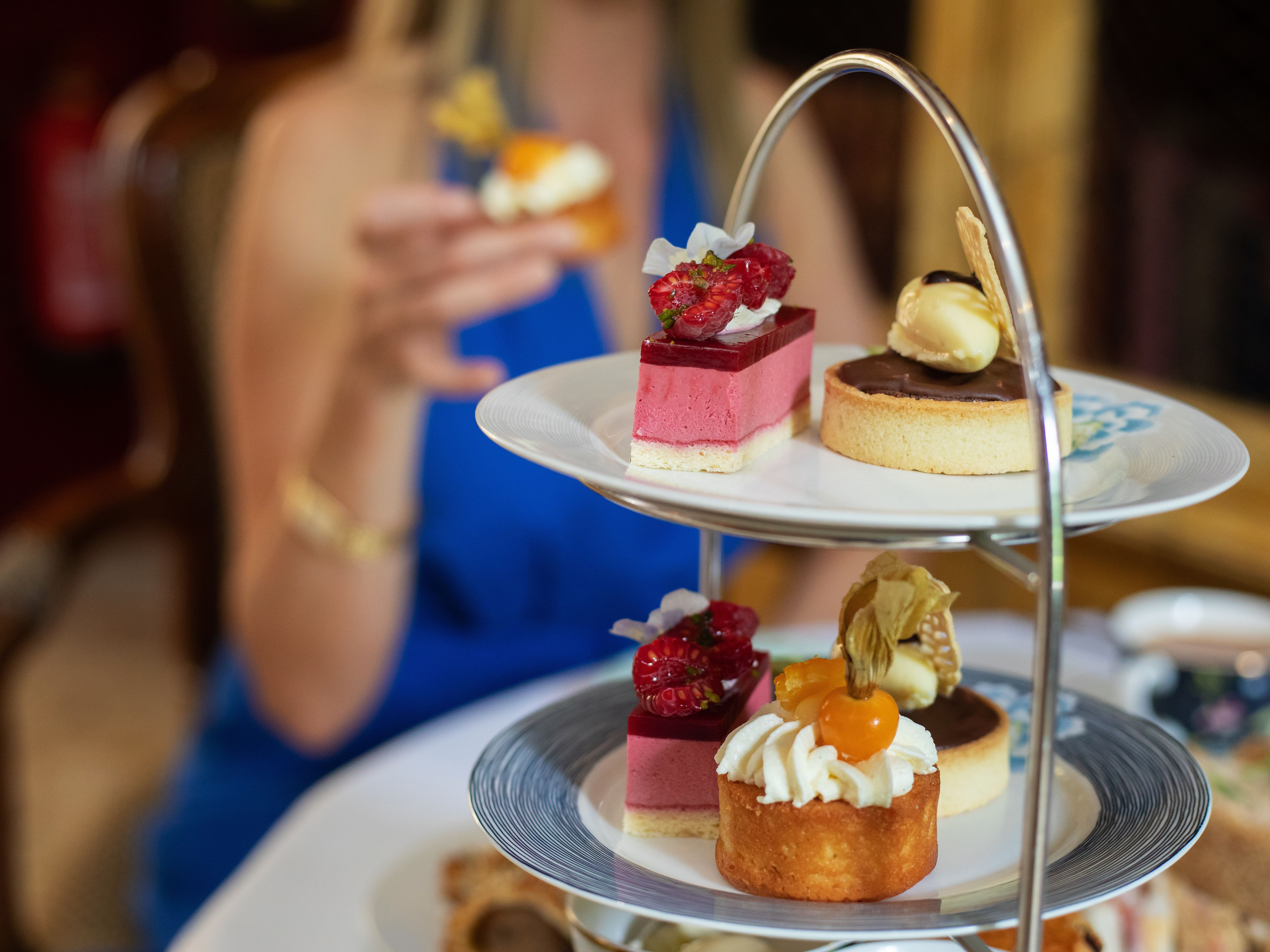 Afternoon tea table with a woman in a blue dress holding a small cake, and a tiered stand filled with colorful desserts and pastries.
