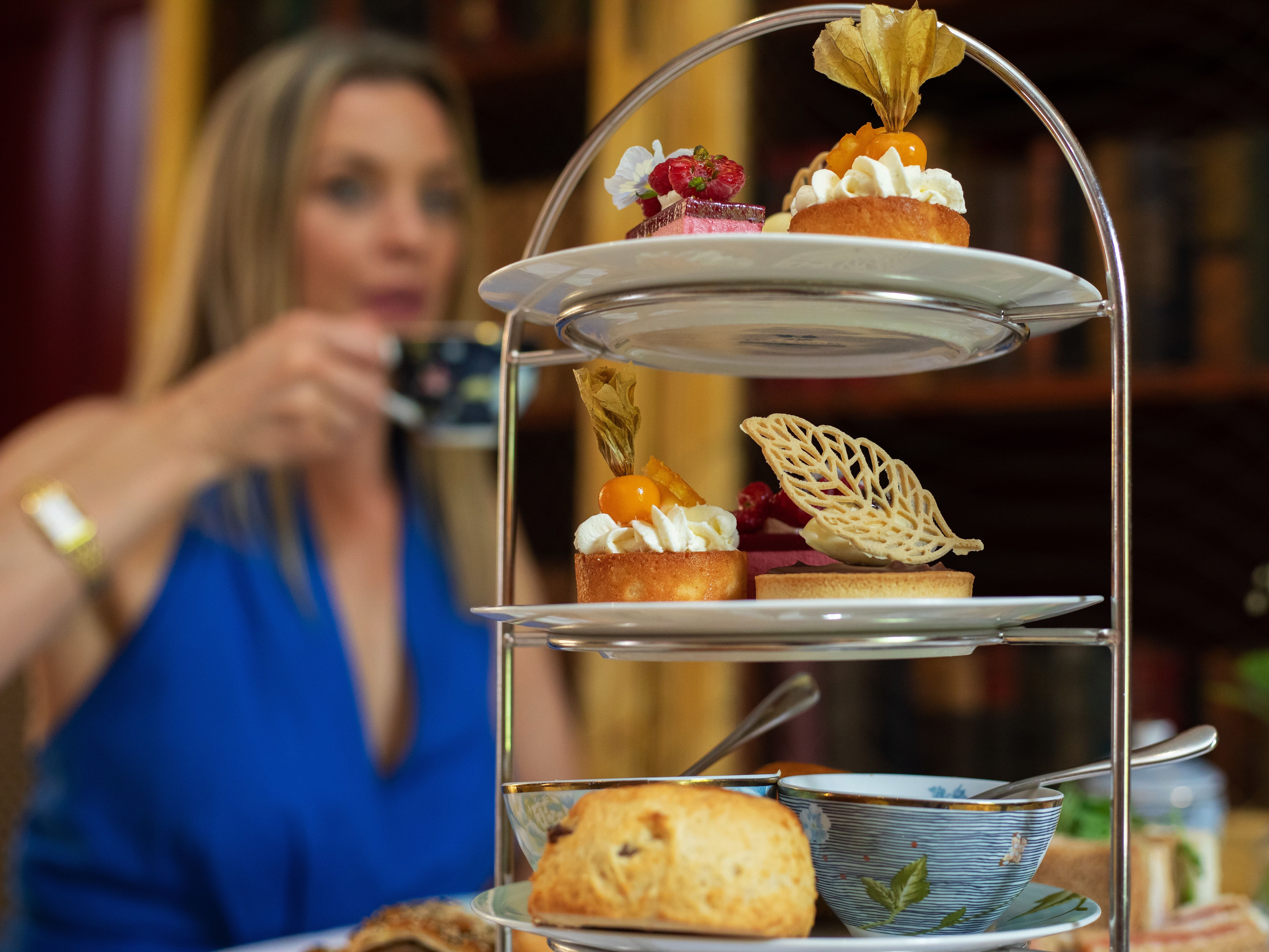 Afternoon tea with pastries and scones on a tiered stand, with a woman in a blue dress holding a teacup in the background.