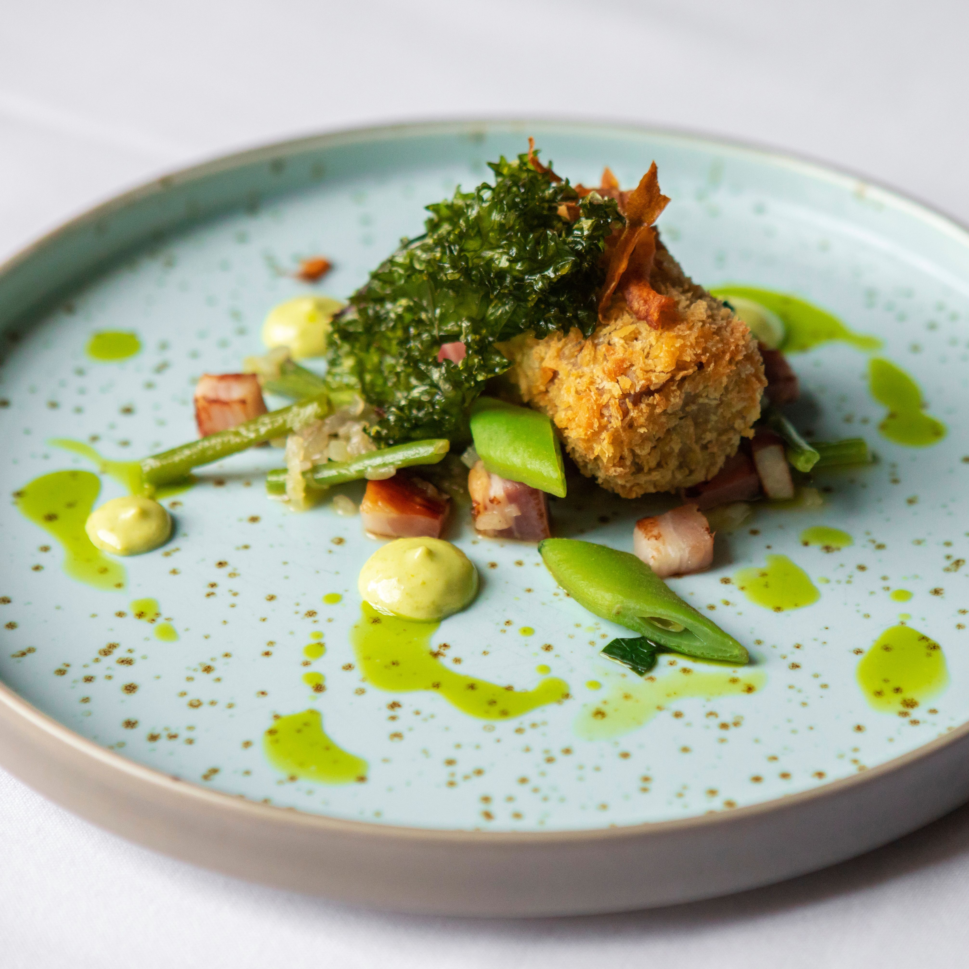A gourmet plated dish featuring a crispy croquette, green vegetables, diced bacon, and herb sauce artfully arranged on a speckled light blue plate.