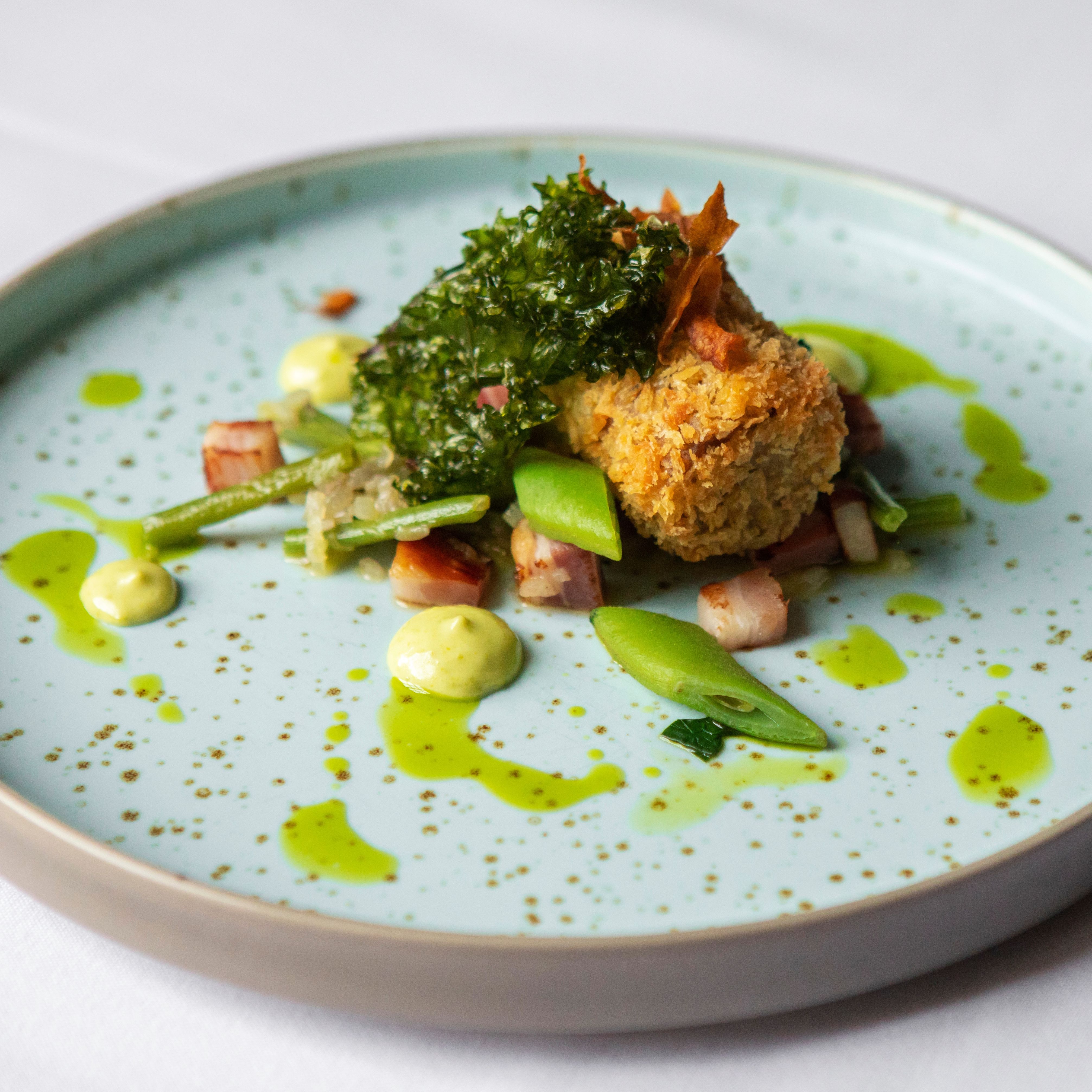 A gourmet plated dish featuring a crispy croquette, green vegetables, diced bacon, and herb sauce artfully arranged on a speckled light blue plate.