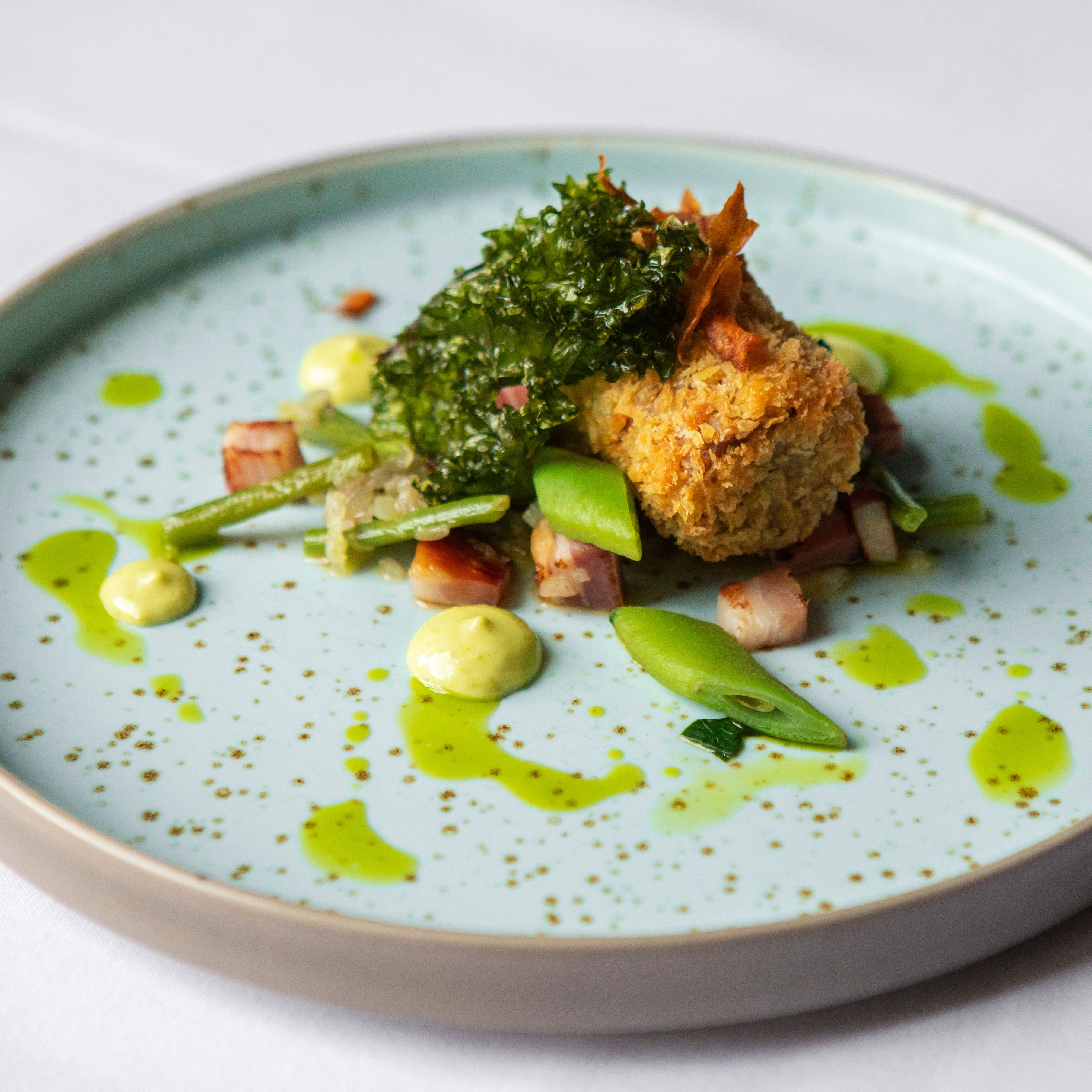 A gourmet plated dish featuring a crispy croquette, green vegetables, diced bacon, and herb sauce artfully arranged on a speckled light blue plate.