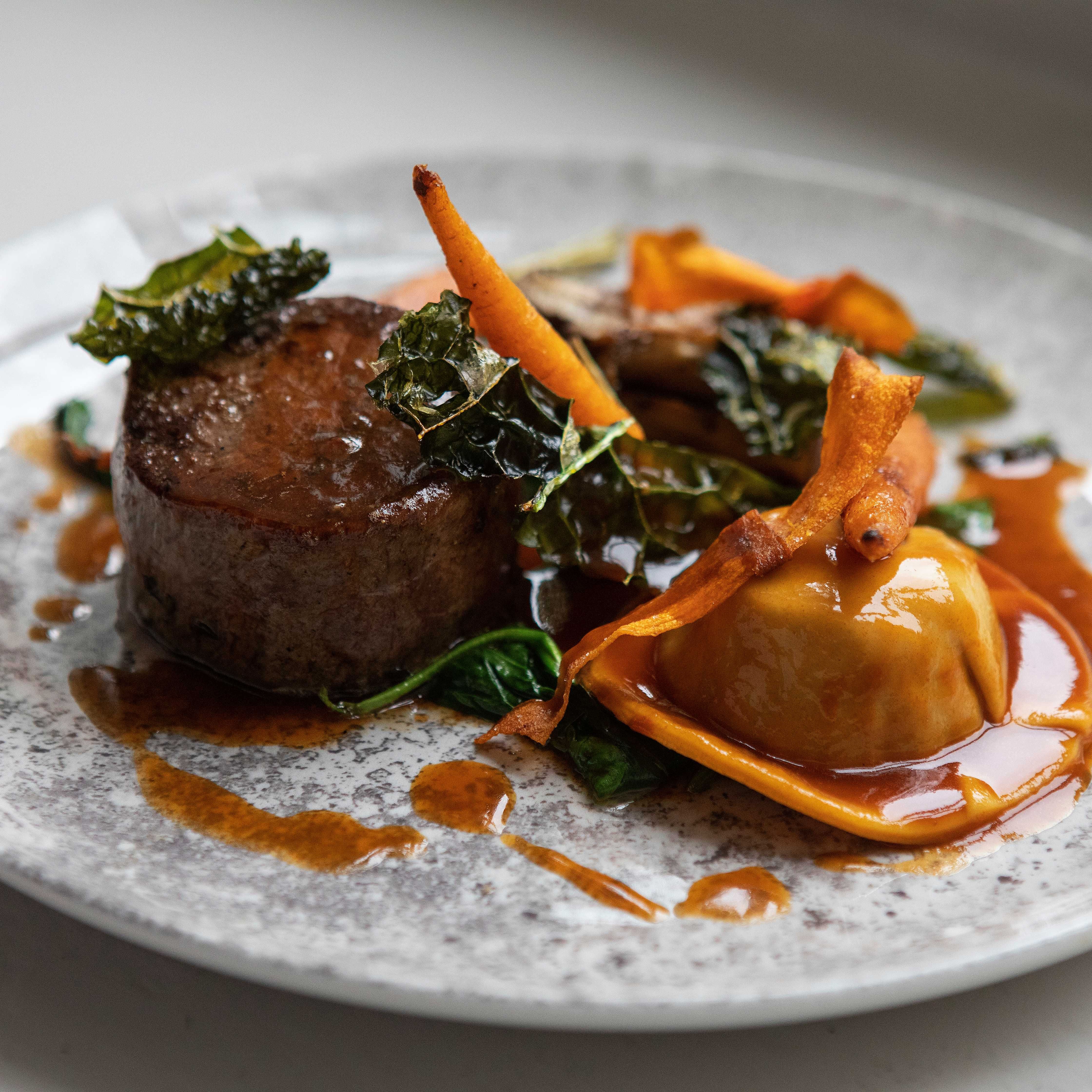 Gourmet plated dish with steak, ravioli, vegetables, and sauce