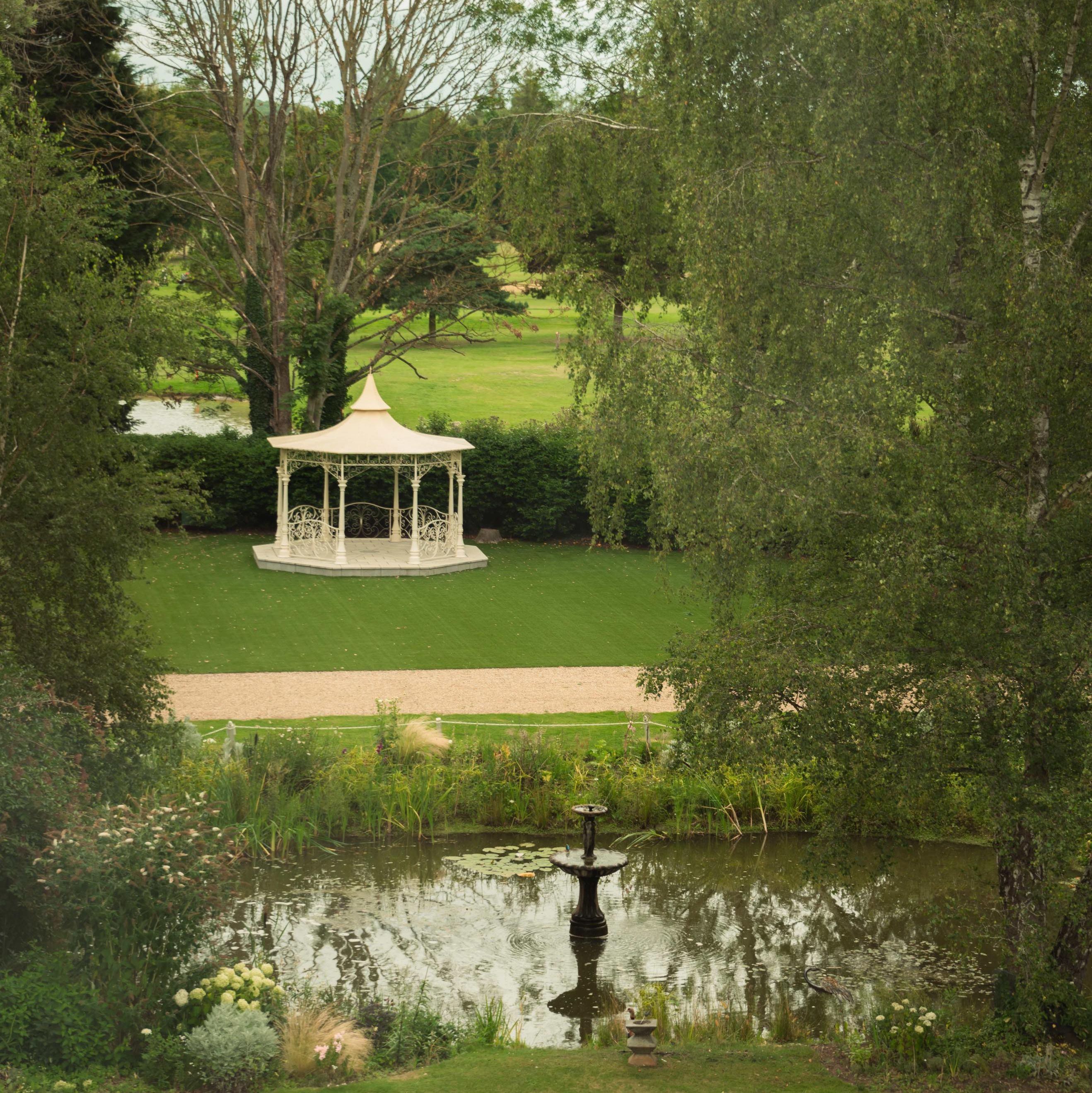 A scenic garden with a white gazebo, a fountain in a pond, and lush greenery.
