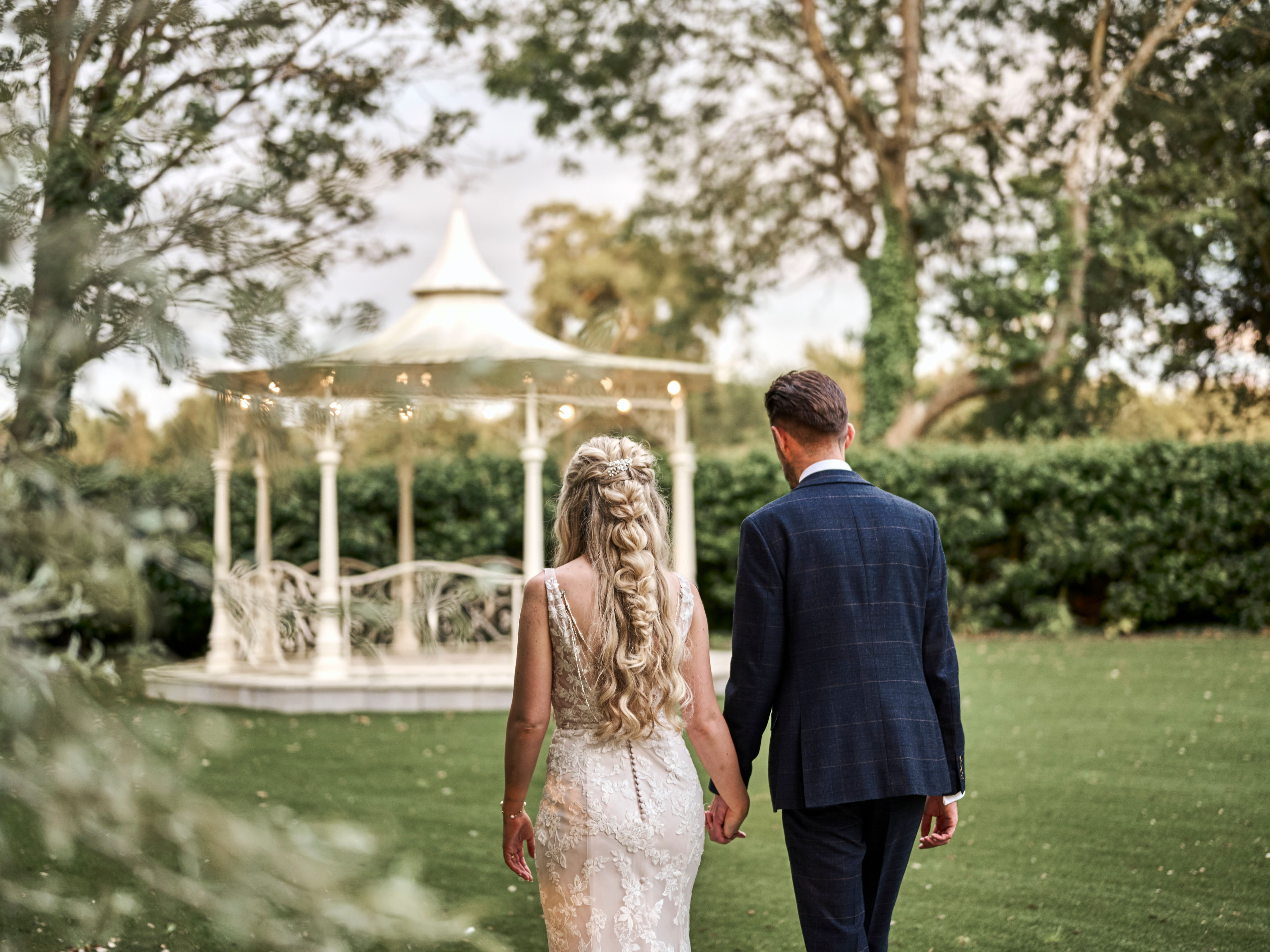 Bride and groom walking hand in hand towards a white gazebo in a garden setting.