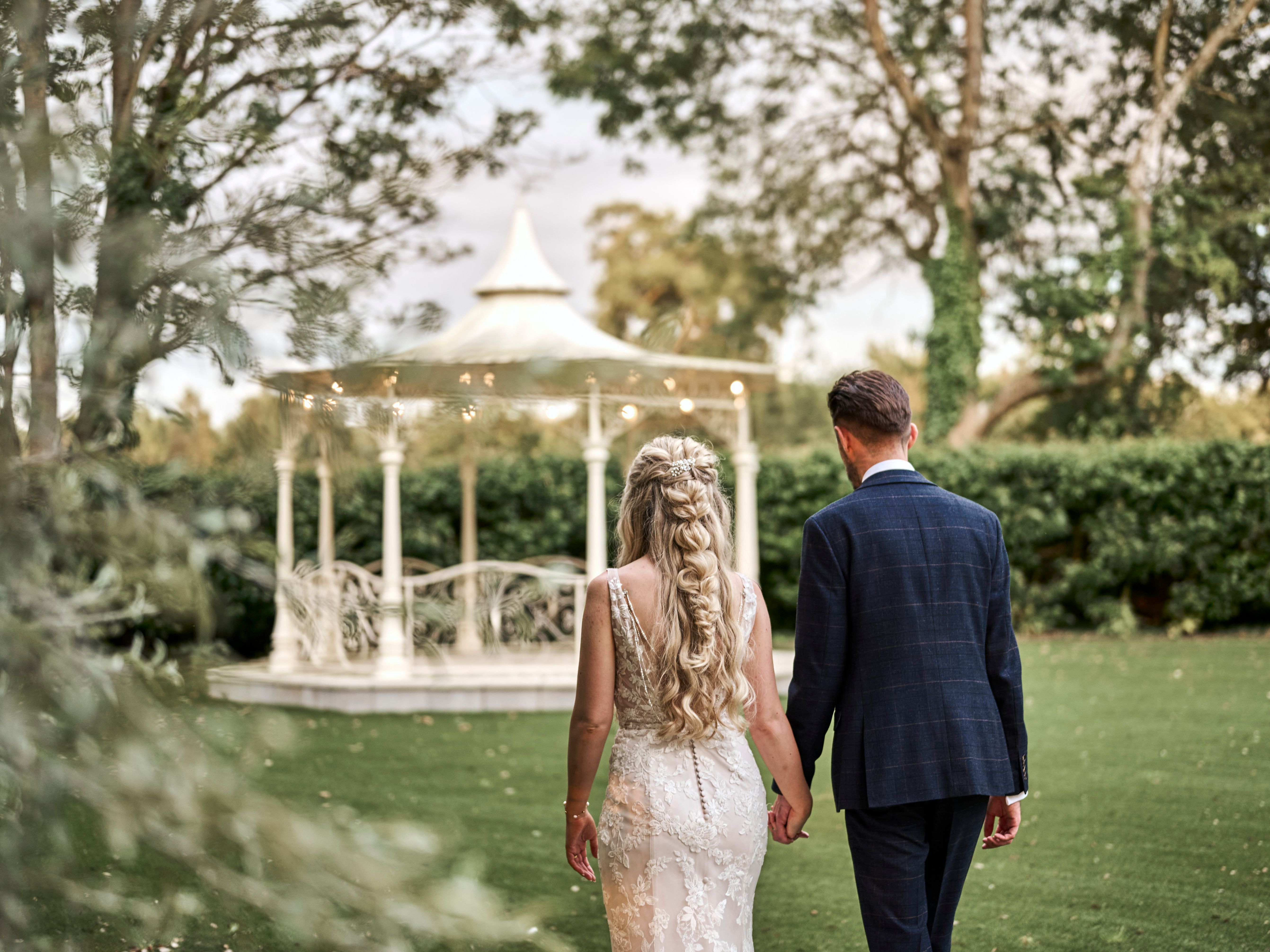 Bride and groom walking hand in hand towards a white gazebo in a garden setting.