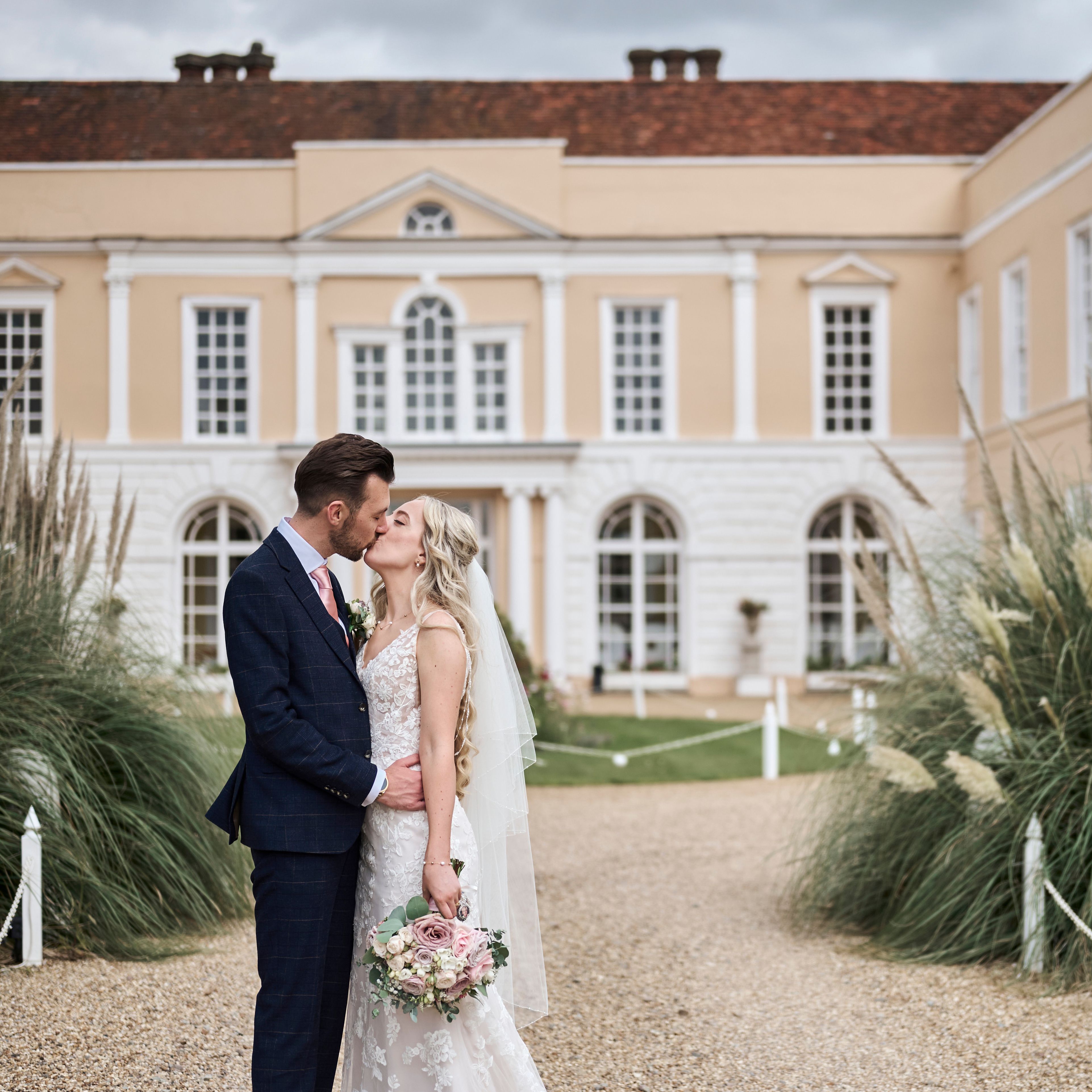 Bride and groom kissing outside a grand historic building with lush greenery around them.