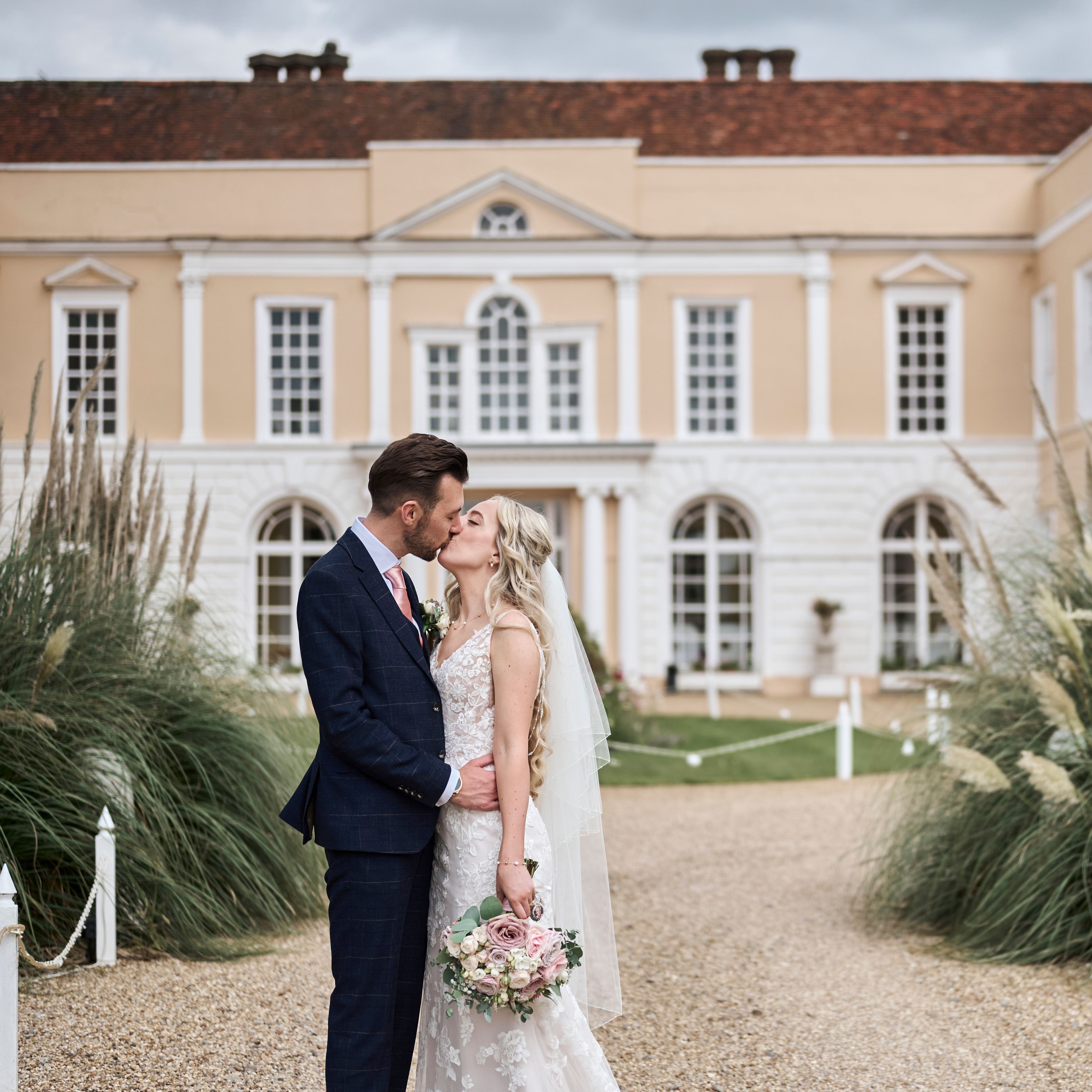 Bride and groom kissing outside a grand historic building with lush greenery around them.