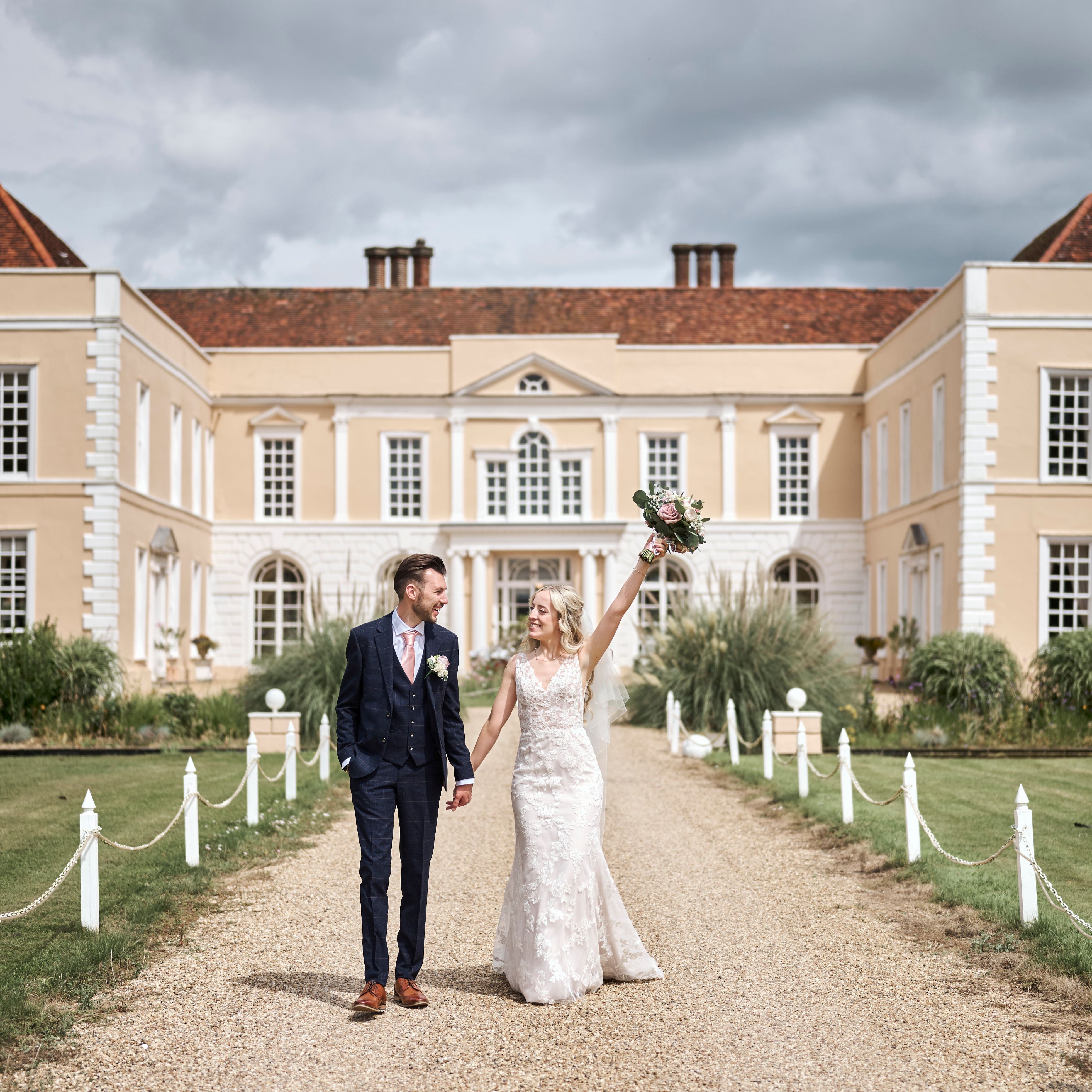 Bride and groom walking hand in hand outside a grand manor house, with the bride raising her bouquet joyfully.