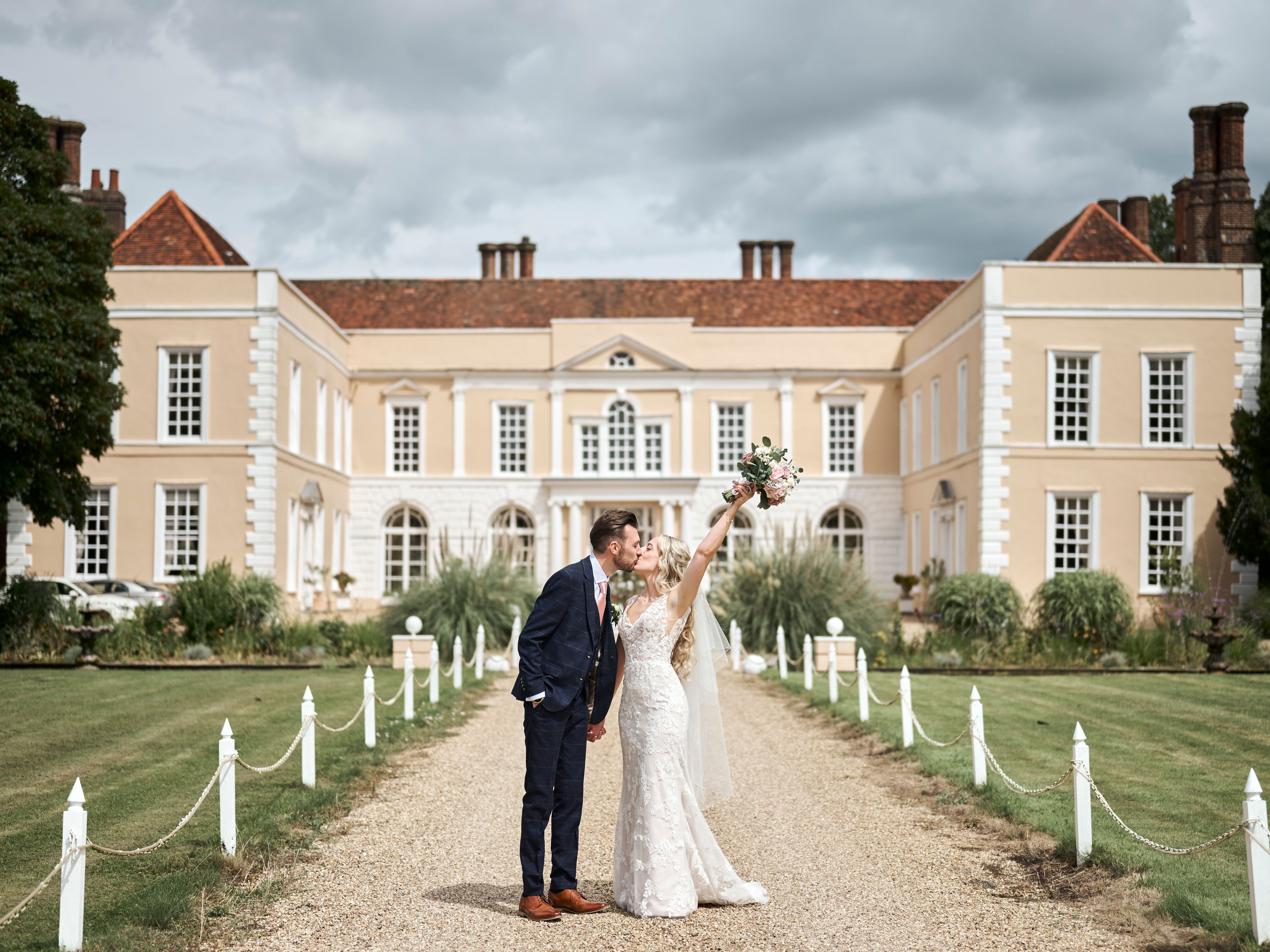 Bride and groom kissing outside Hintlesham Hall, with the bride raising her bouquet in celebration.