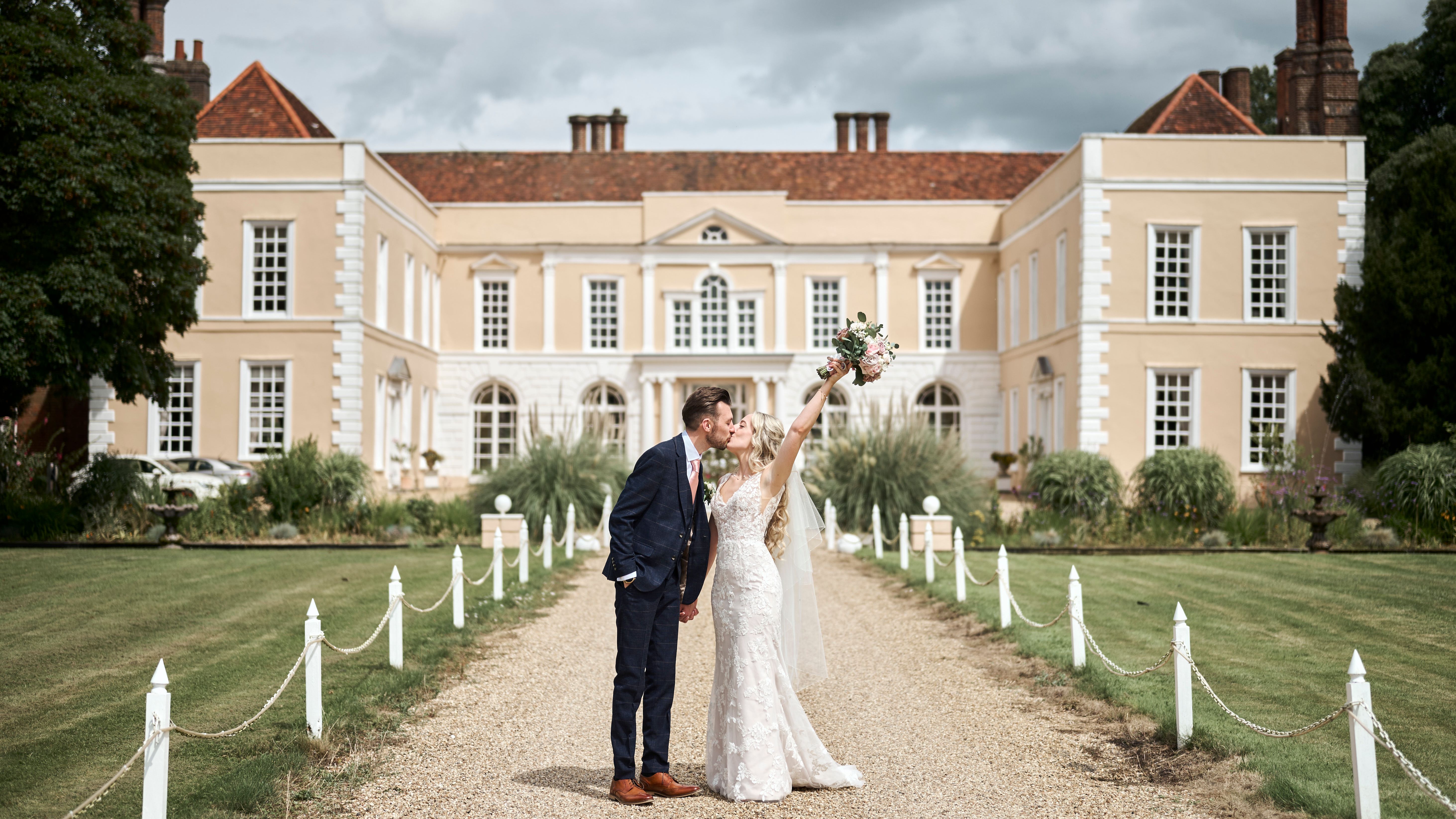 Bride and groom kissing outside Hintlesham Hall, with the bride raising her bouquet in celebration.