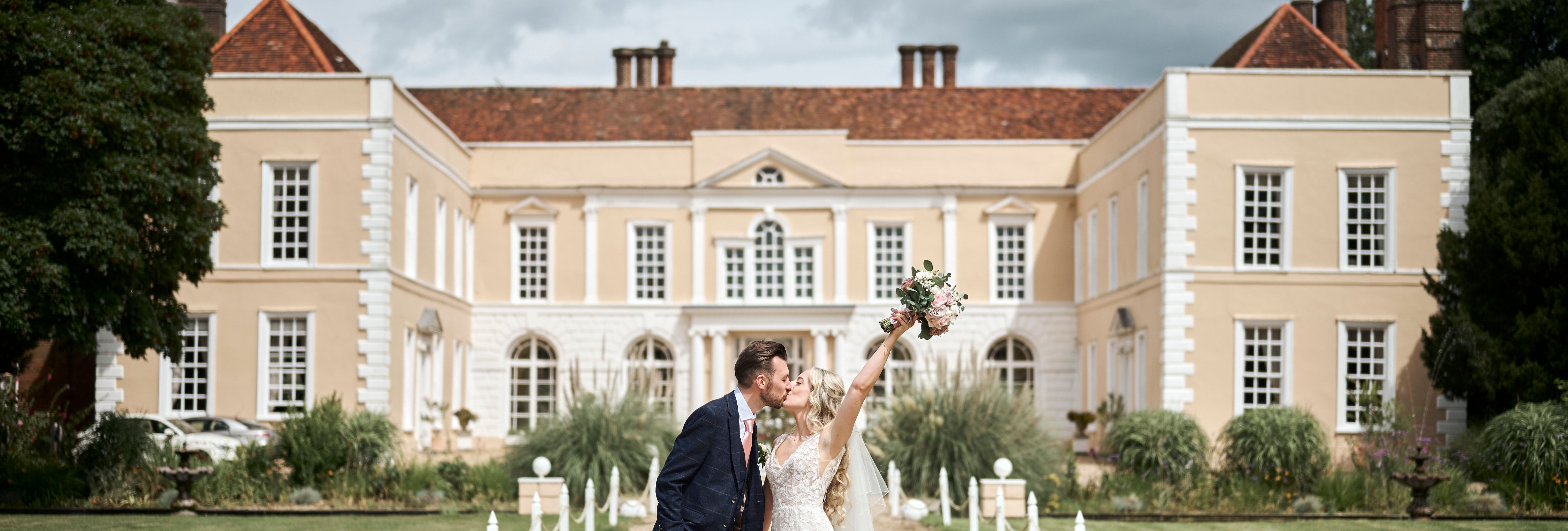 Bride and groom kissing outside Hintlesham Hall, with the bride raising her bouquet in celebration.