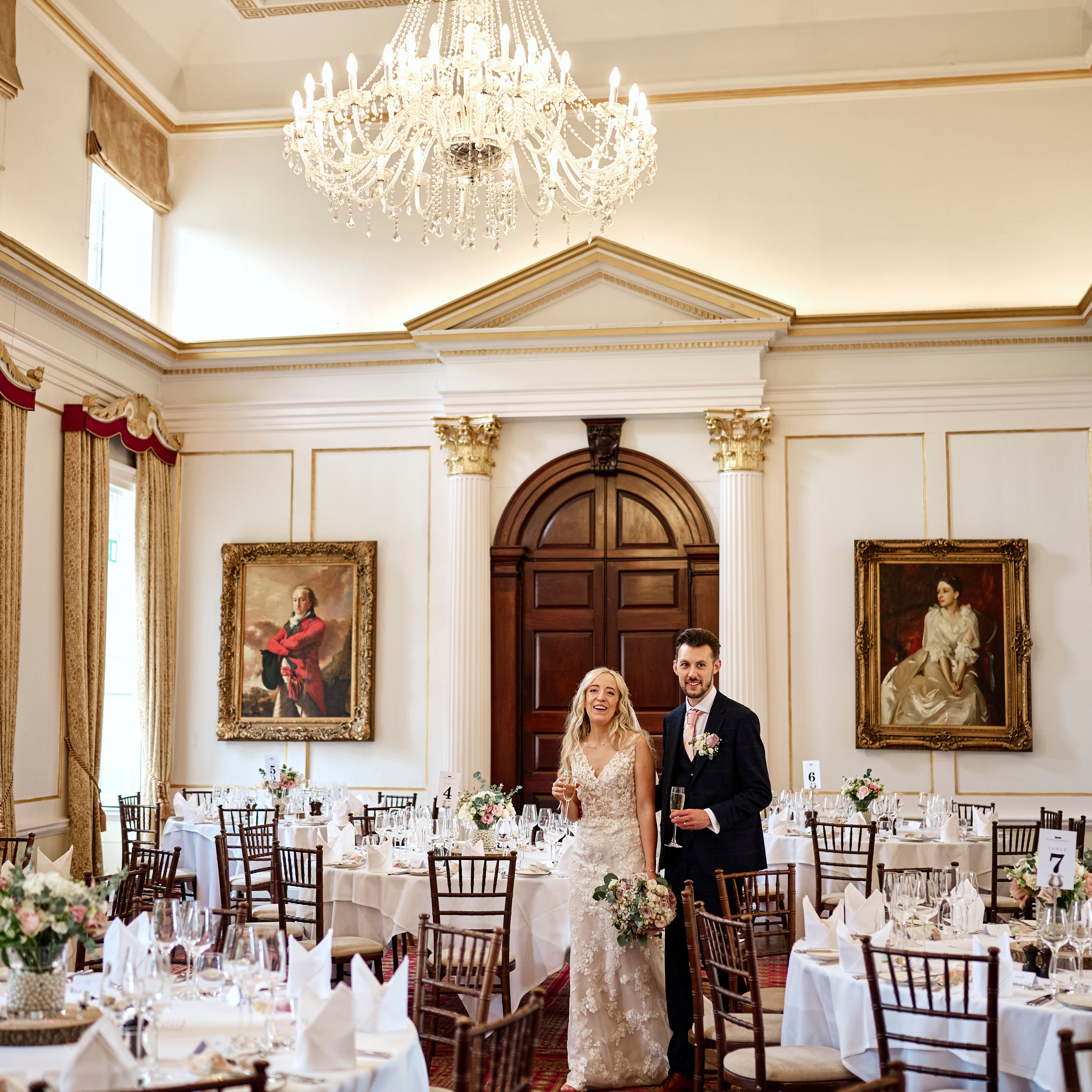 A bride and groom stand together in an elegant, chandelier-lit dining room decorated for a wedding reception.