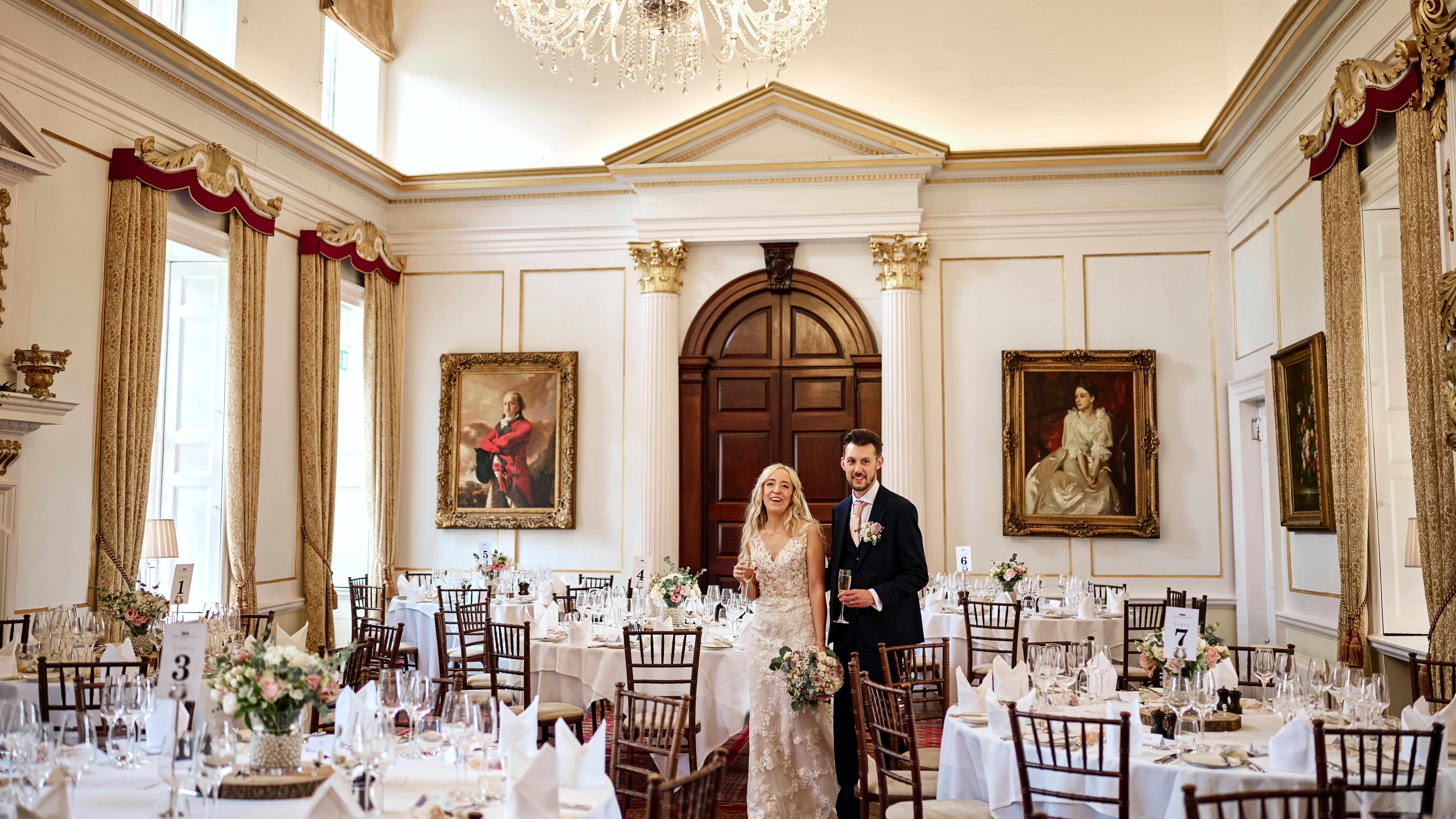 A bride and groom stand together in an elegant, chandelier-lit dining room decorated for a wedding reception.