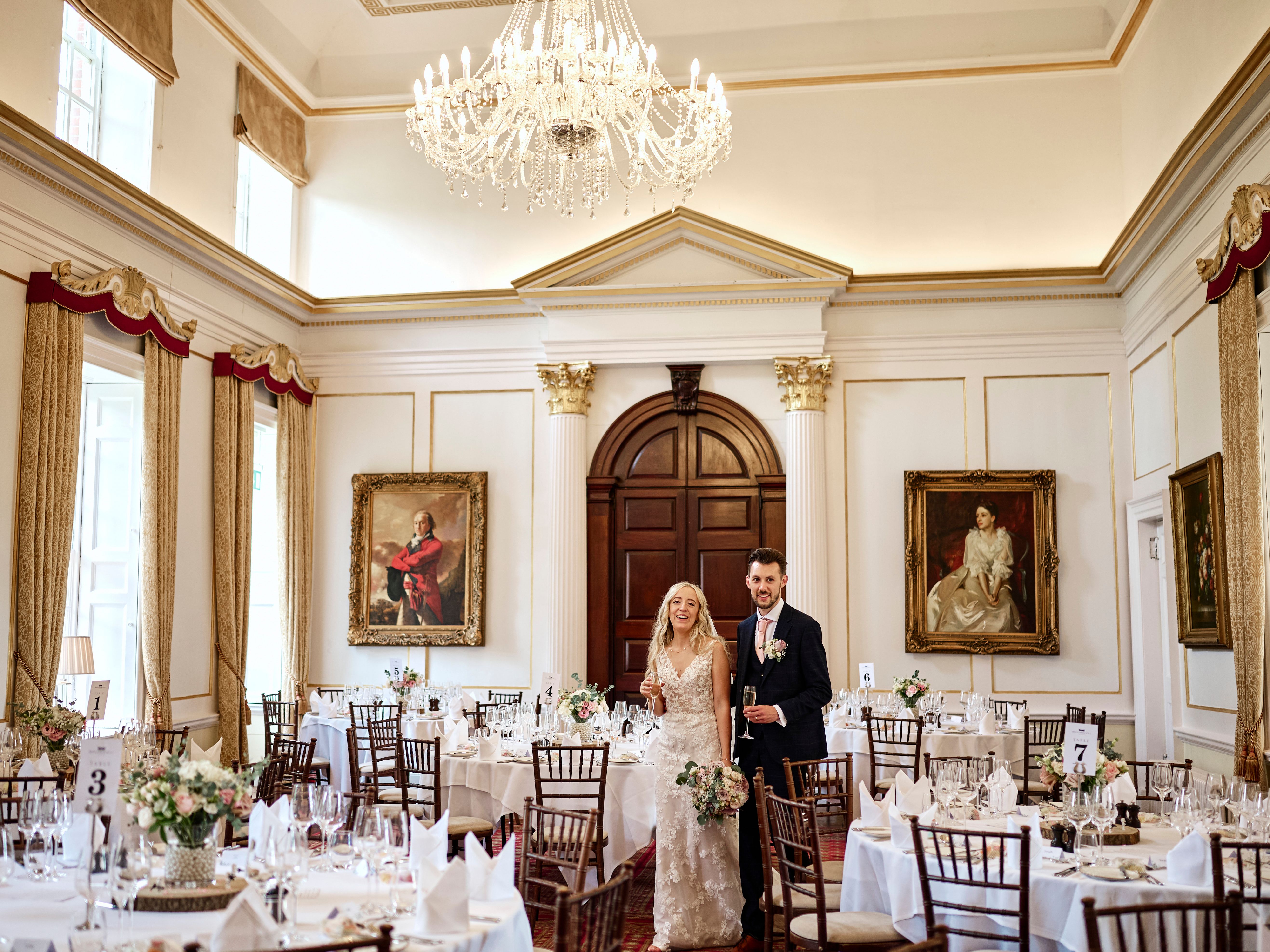 A bride and groom stand together in an elegant, chandelier-lit dining room decorated for a wedding reception.