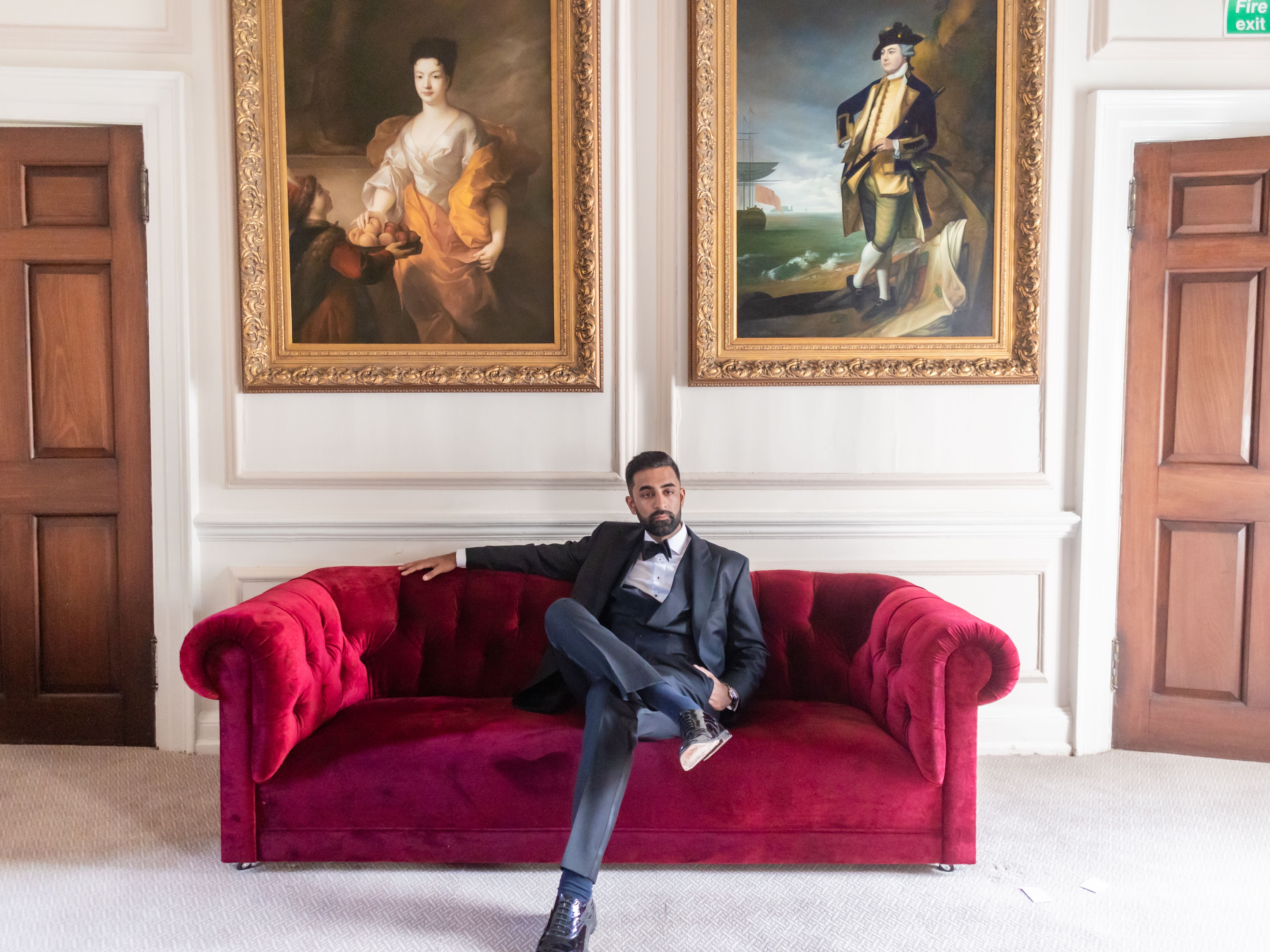 Man in formal attire sitting on a red velvet sofa beneath two large classical paintings in an elegant room.