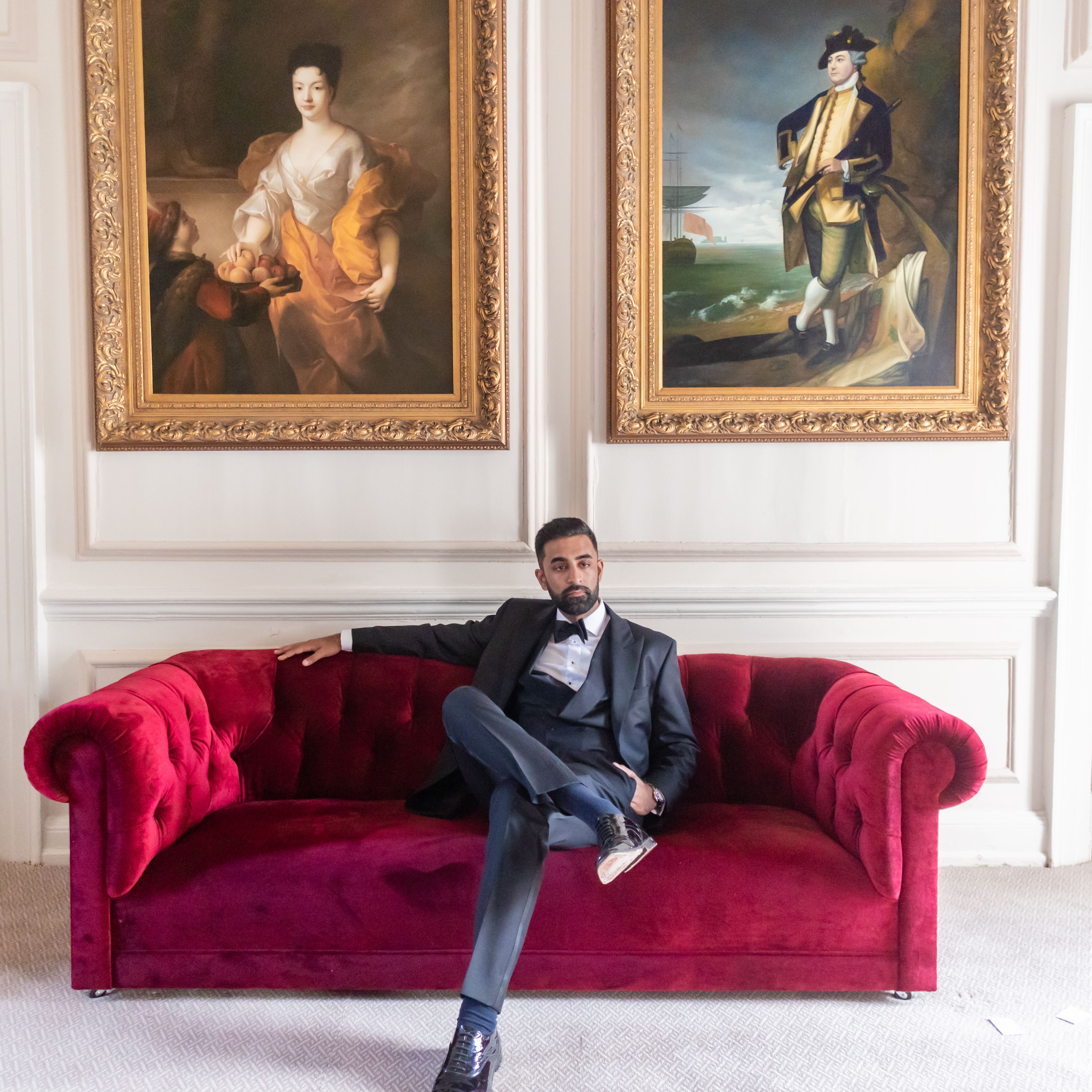 Man in formal attire sitting on a red velvet sofa beneath two large classical paintings in an elegant room.