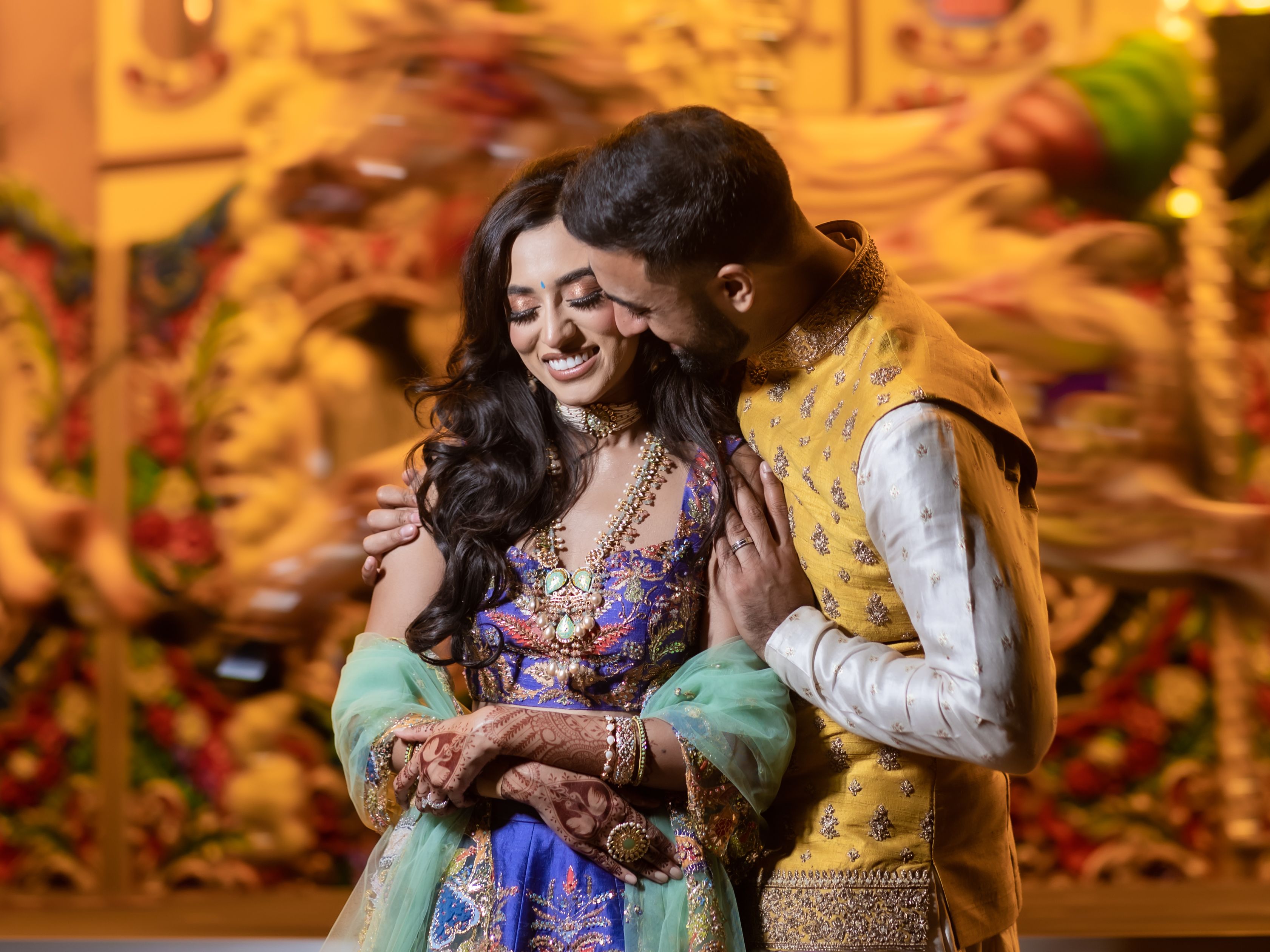 A couple in traditional Indian attire embracing and smiling in front of a brightly lit carousel.