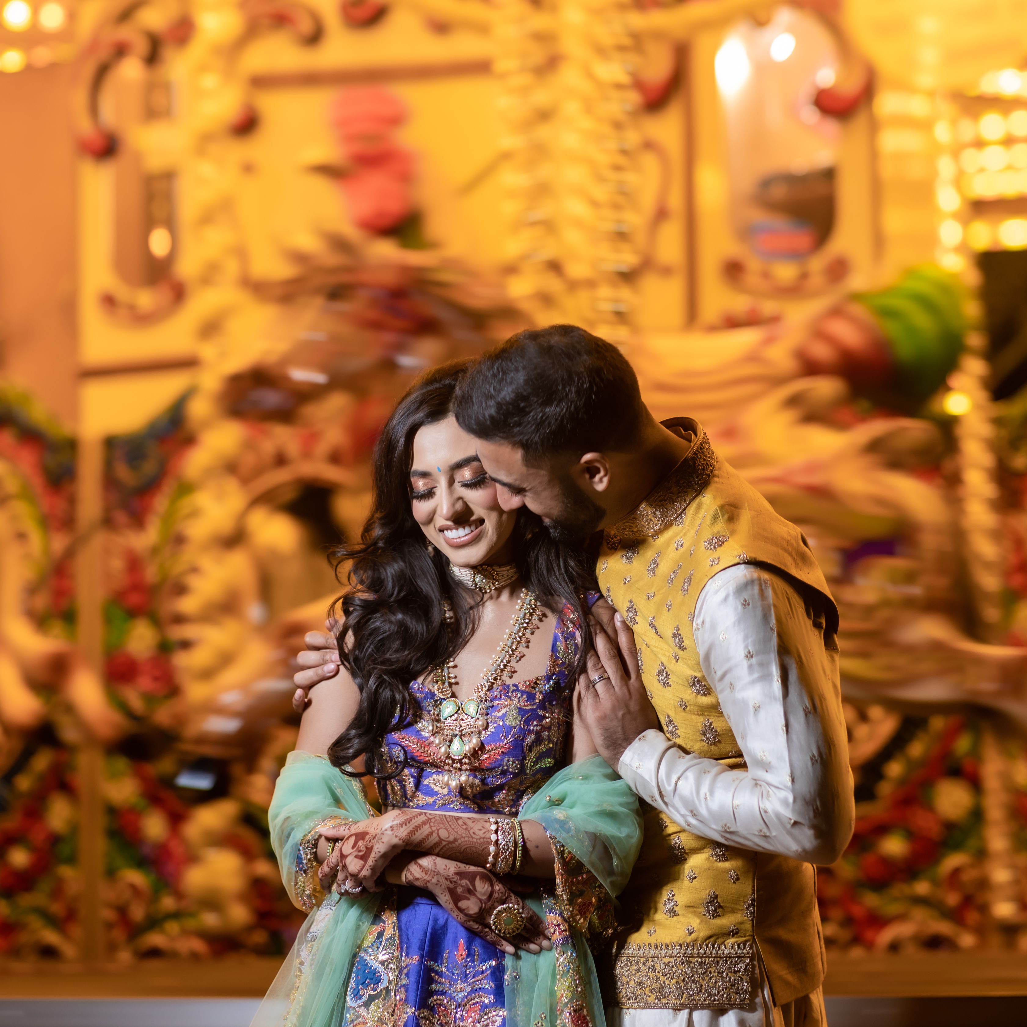 A couple in traditional Indian attire embracing and smiling in front of a brightly lit carousel.