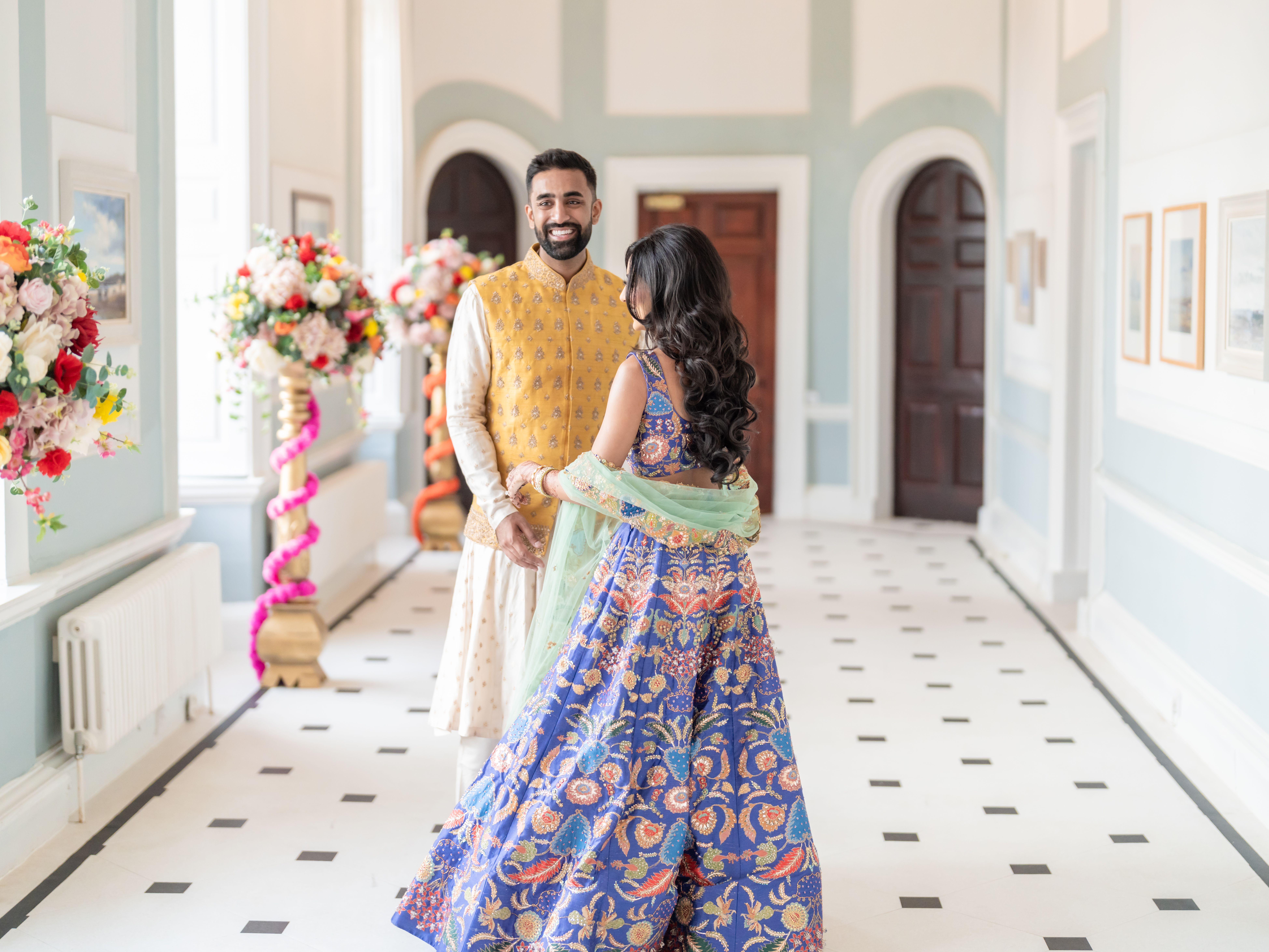 A couple in traditional South Asian attire smiles at each other in a bright, elegantly decorated hallway with large flower arrangements.