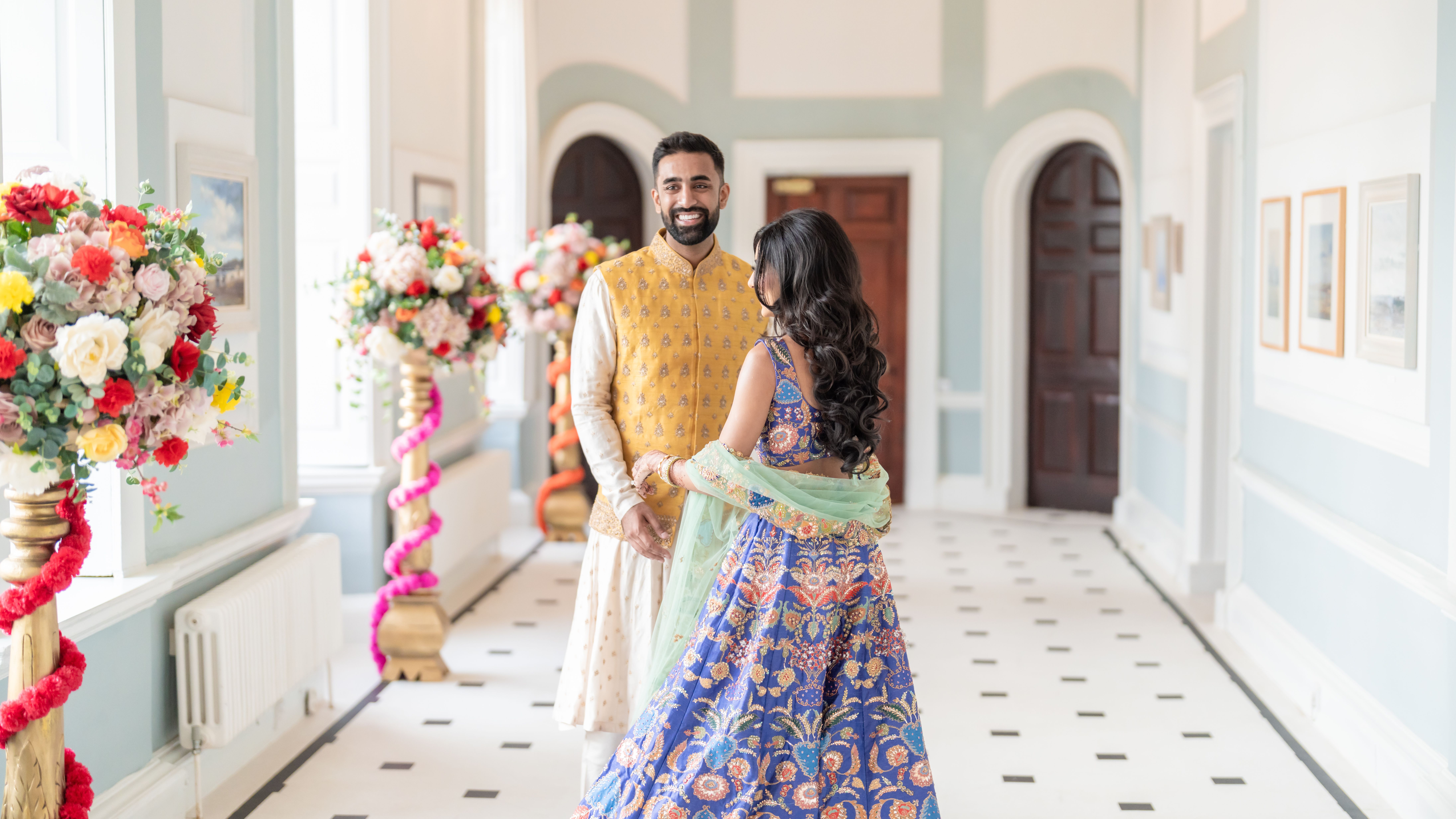 A couple in traditional South Asian attire smiles at each other in a bright, elegantly decorated hallway with large flower arrangements.
