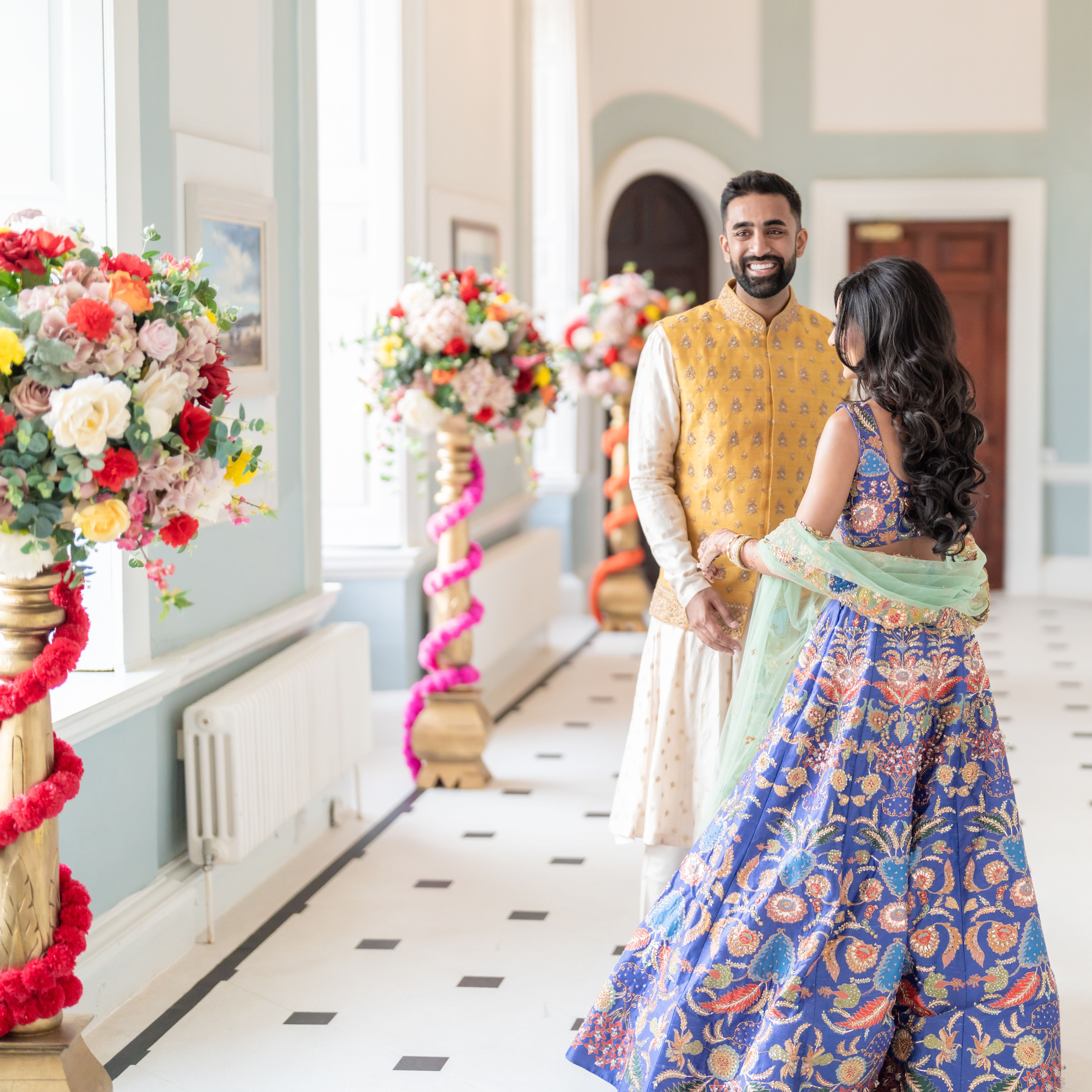 A couple in traditional South Asian attire smiles at each other in a bright, elegantly decorated hallway with large flower arrangements.