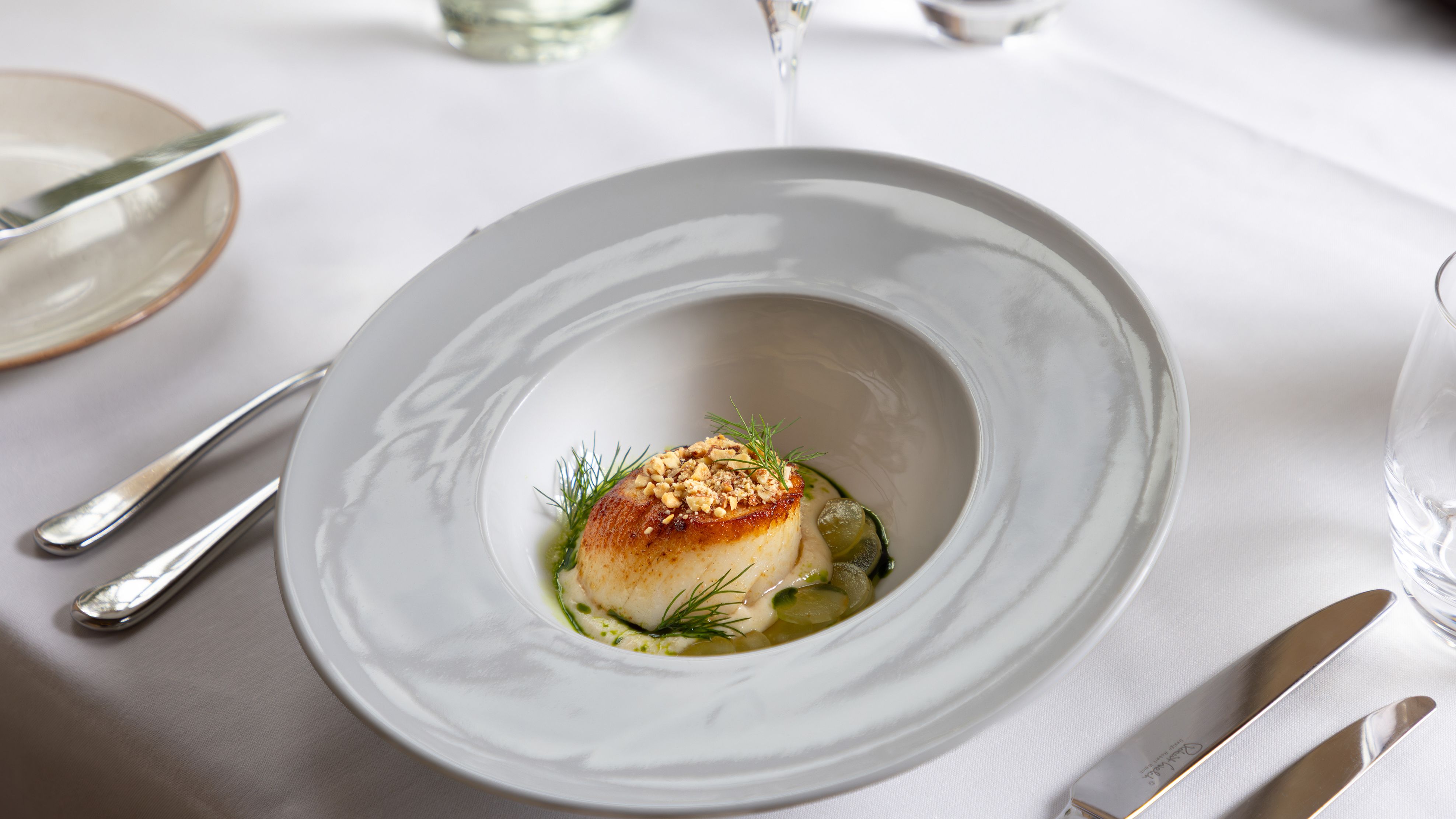 A gourmet dish with a seared scallop on a white plate, garnished with herbs and crumbled nuts, served at a fine dining restaurant.
