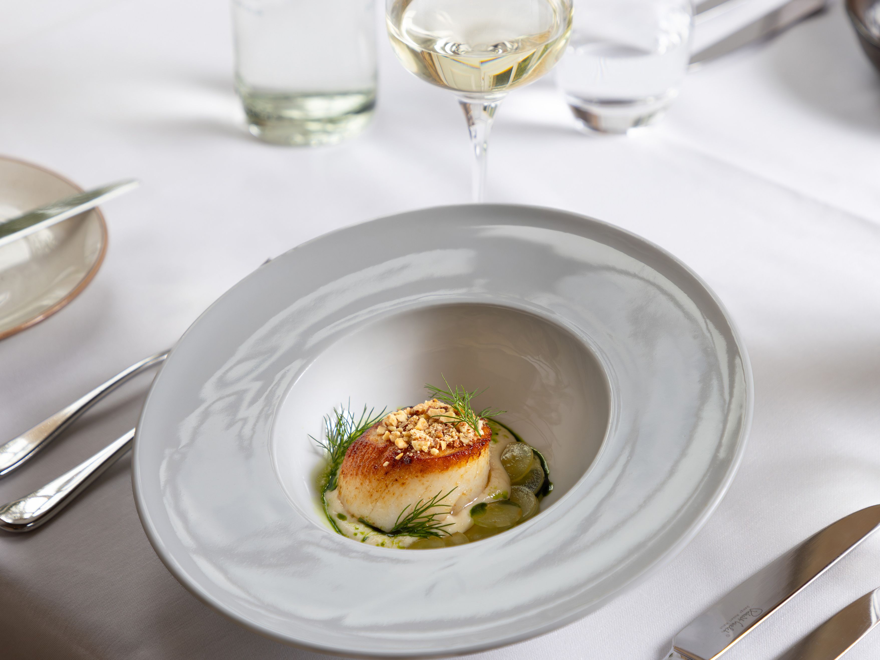 A gourmet dish with a seared scallop on a white plate, garnished with herbs and crumbled nuts, served at a fine dining restaurant.