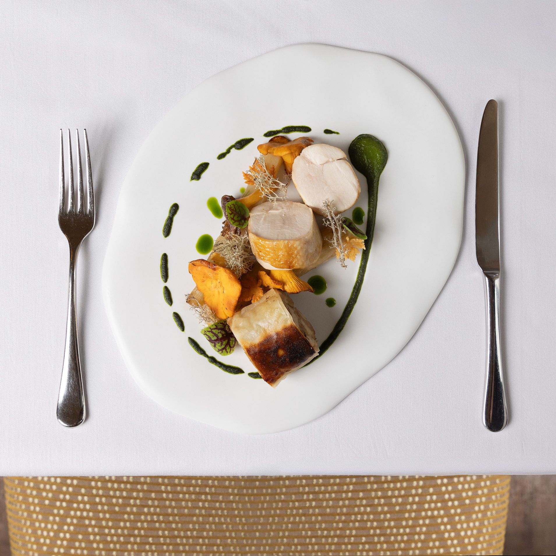 Gourmet plated dish with chicken, mushrooms, and green sauce on a white cloth table setting