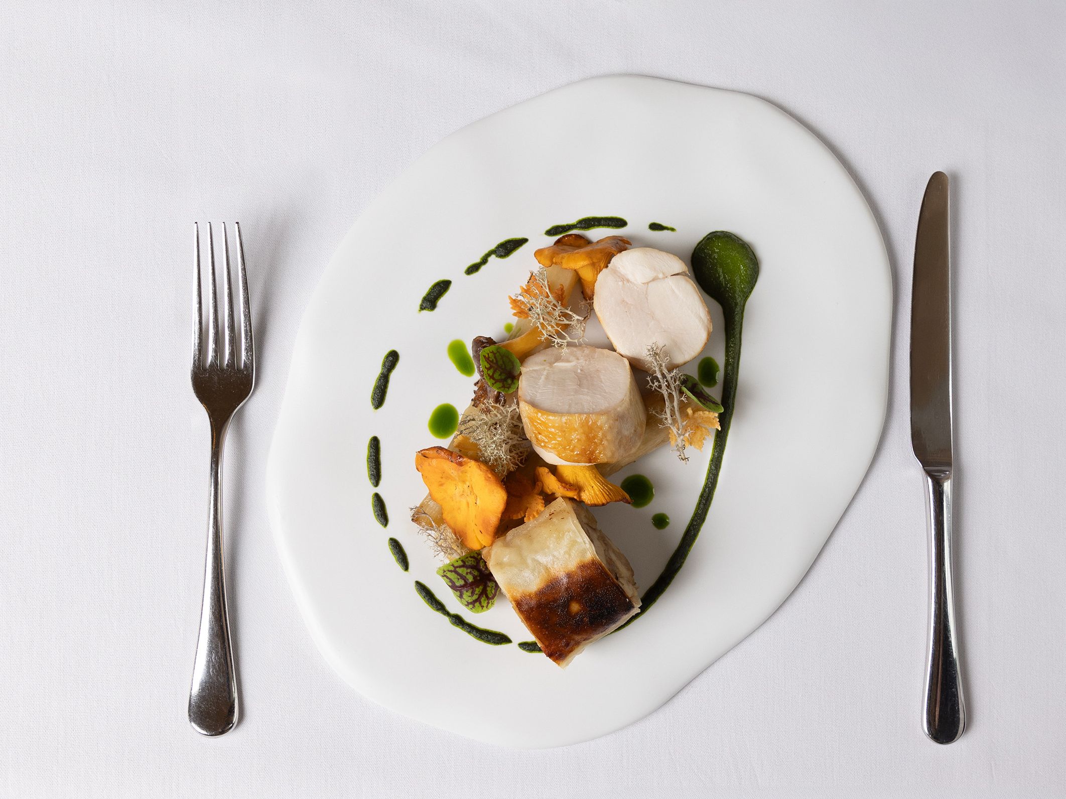 Gourmet plated dish with chicken, mushrooms, and green sauce on a white cloth table setting