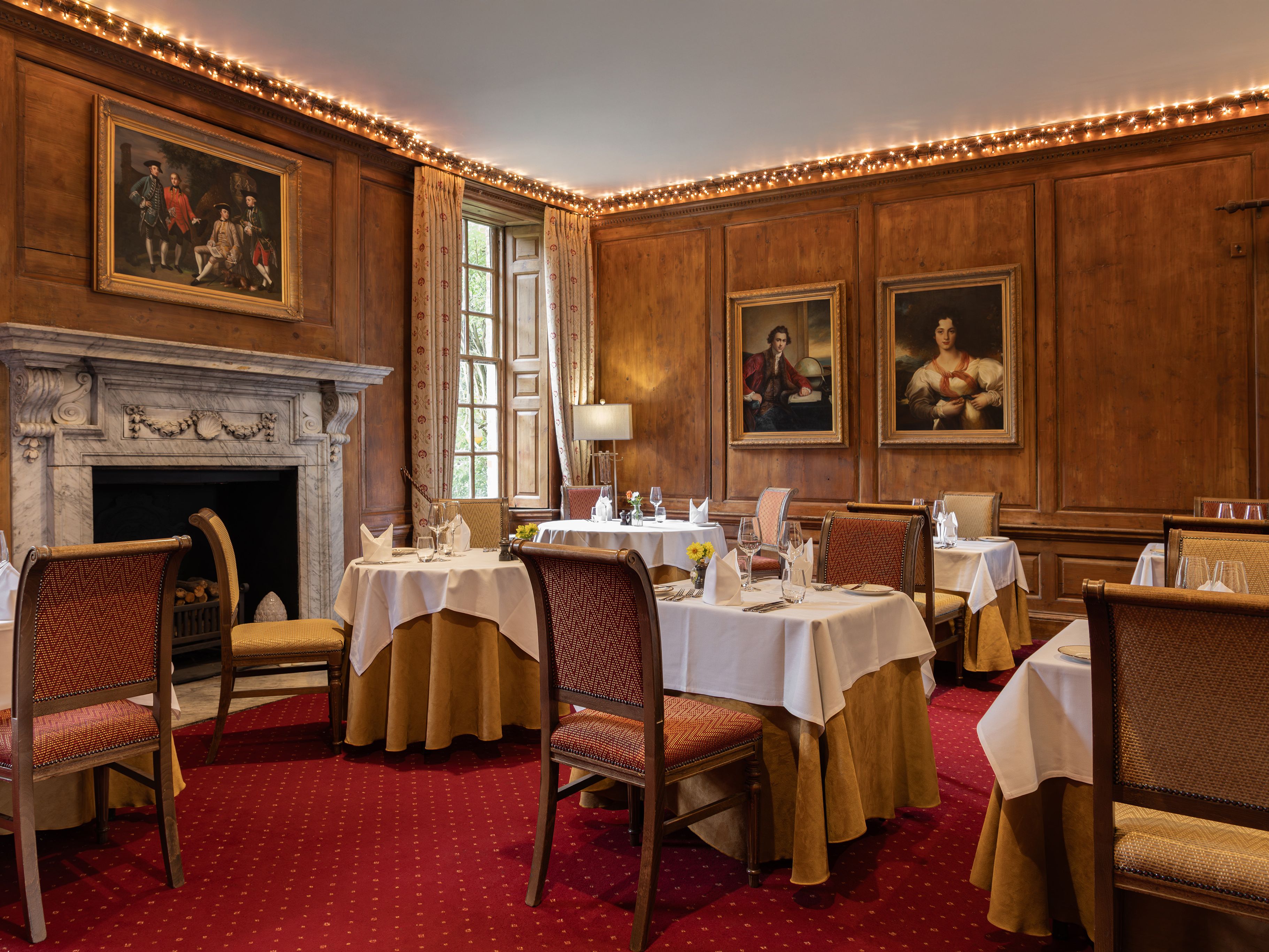 Elegant restaurant with wooden paneled walls, classic paintings, round tables with white tablecloths, and a marble fireplace.