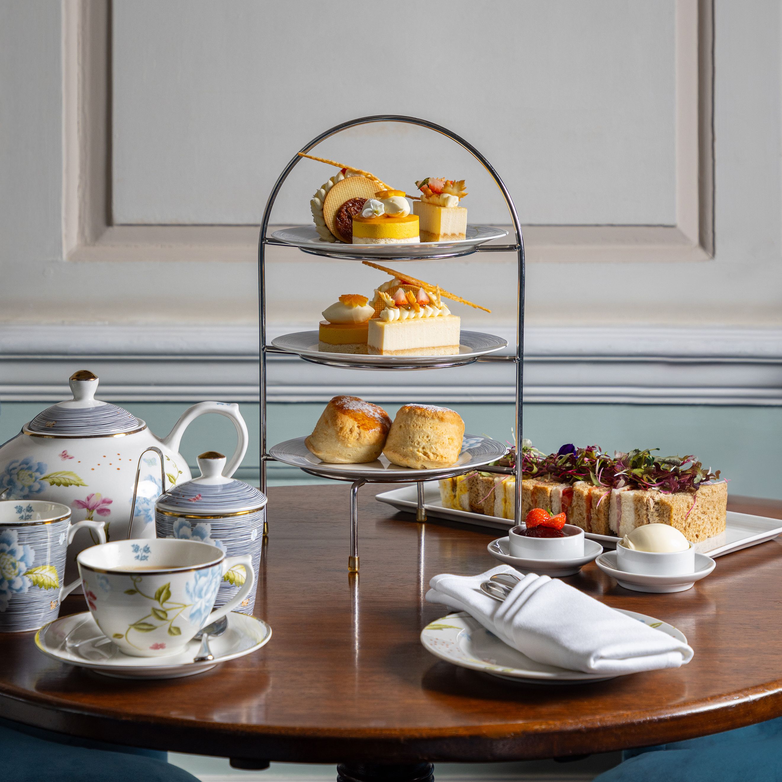 Three-tier afternoon tea stand with pastries, scones, and sandwiches on a wooden table, accompanied by a floral teapot and teacups.
