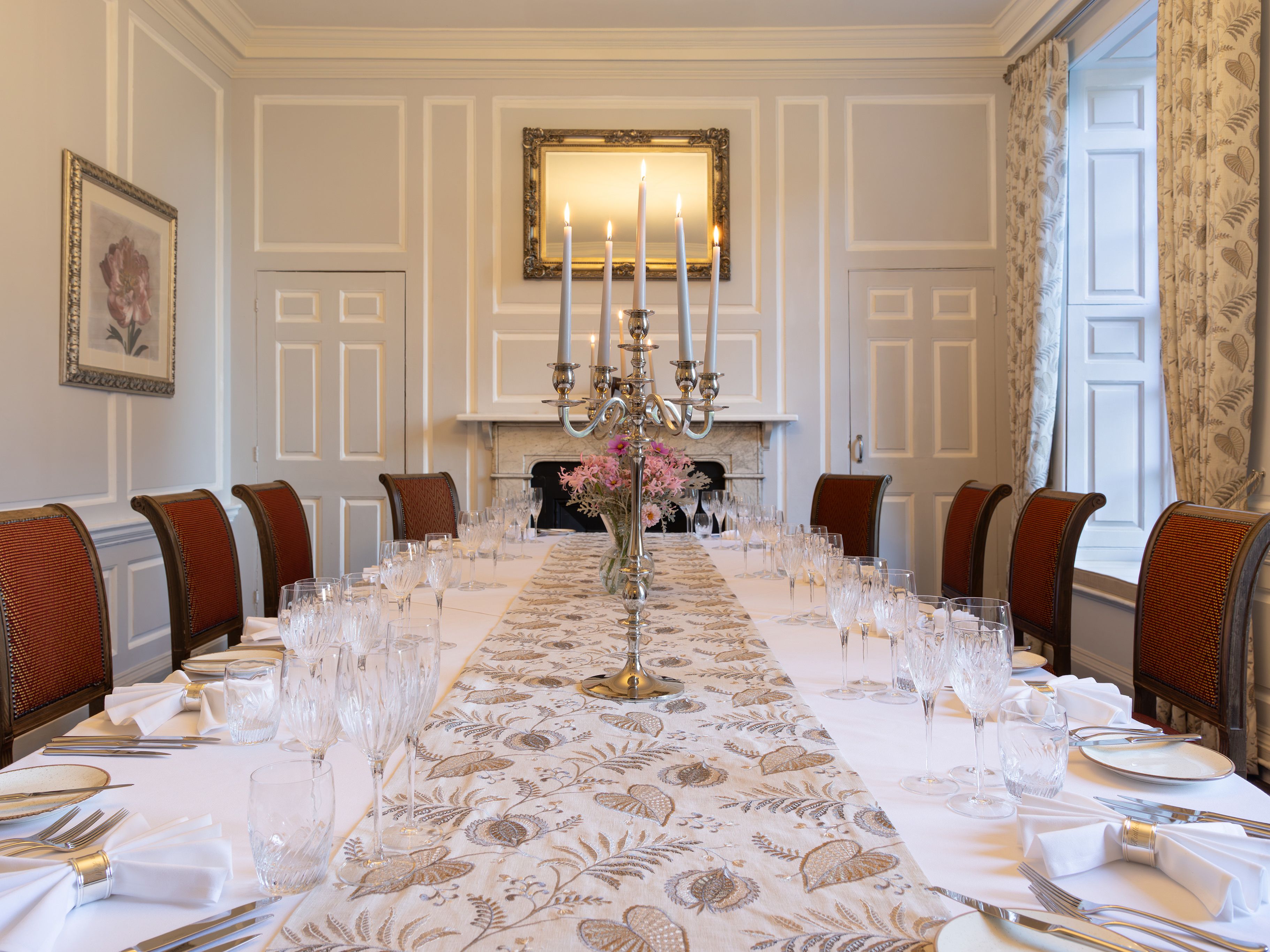 Elegant dining room with a long table set for a formal meal, featuring red chairs, white tablecloth, glassware, and a silver candelabra centerpiece.