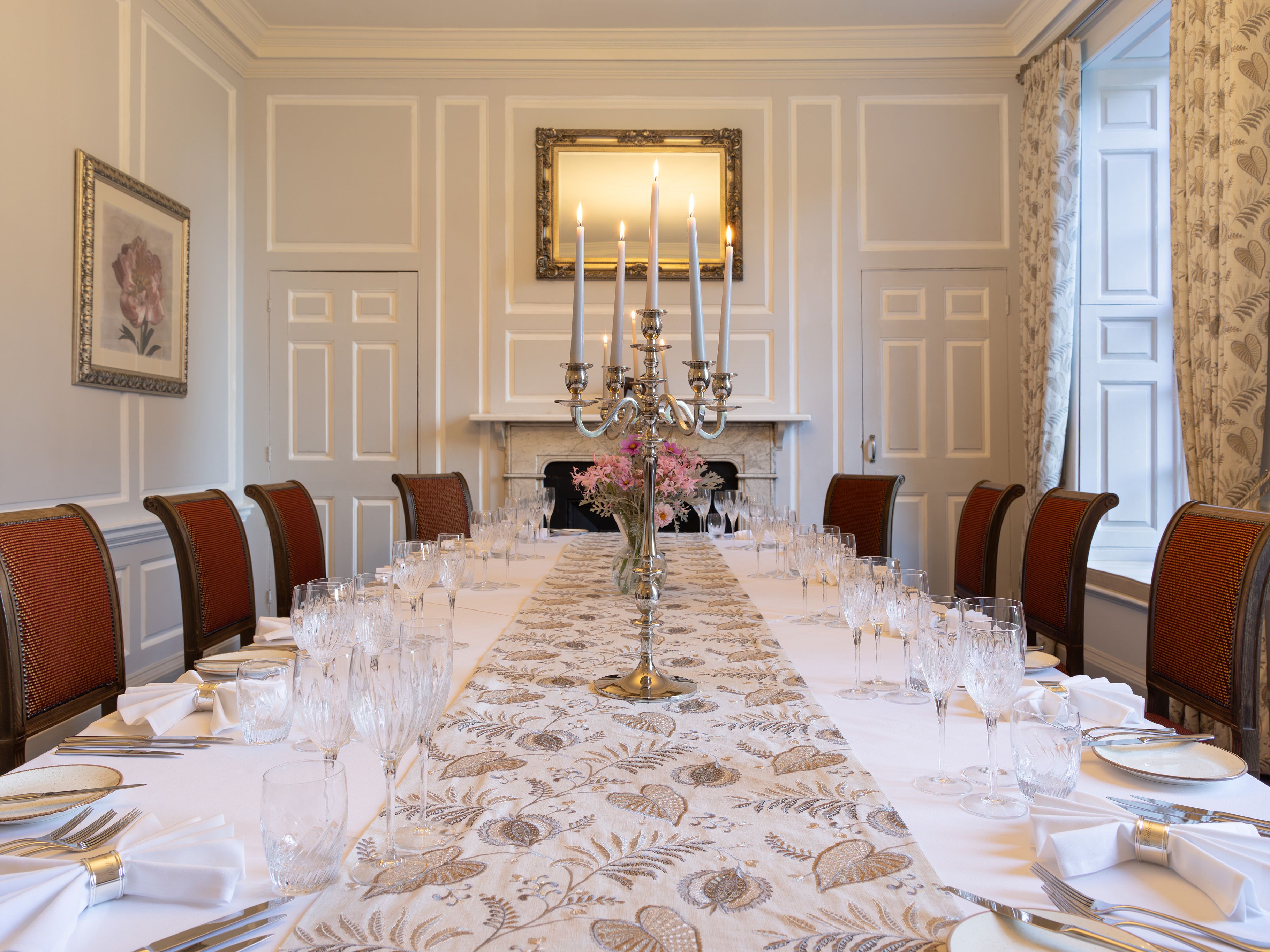 Elegant dining room with a long table set for a formal meal, featuring red chairs, white tablecloth, glassware, and a silver candelabra centerpiece.