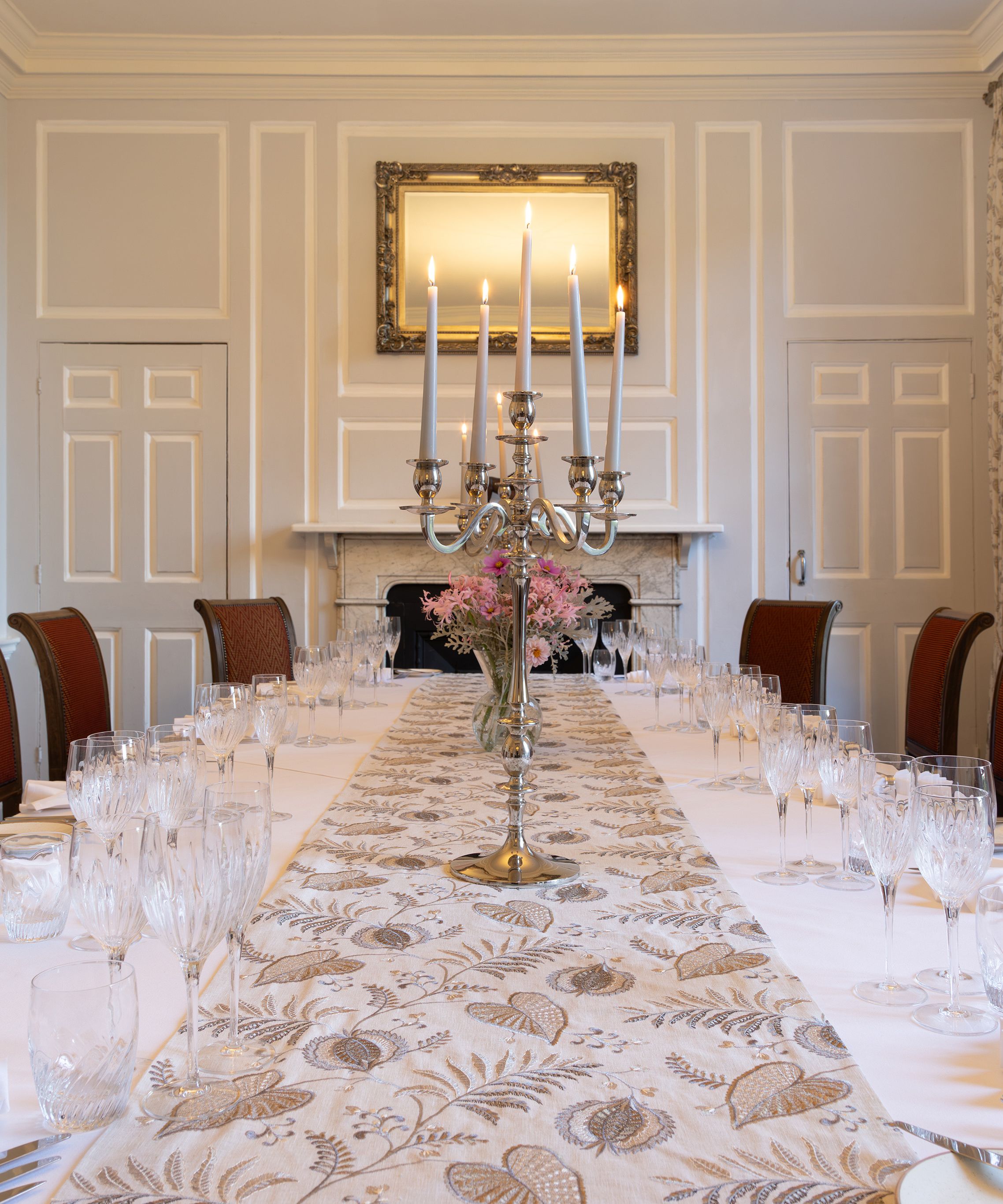 Elegant dining room with a long table set for a formal meal, featuring red chairs, white tablecloth, glassware, and a silver candelabra centerpiece.