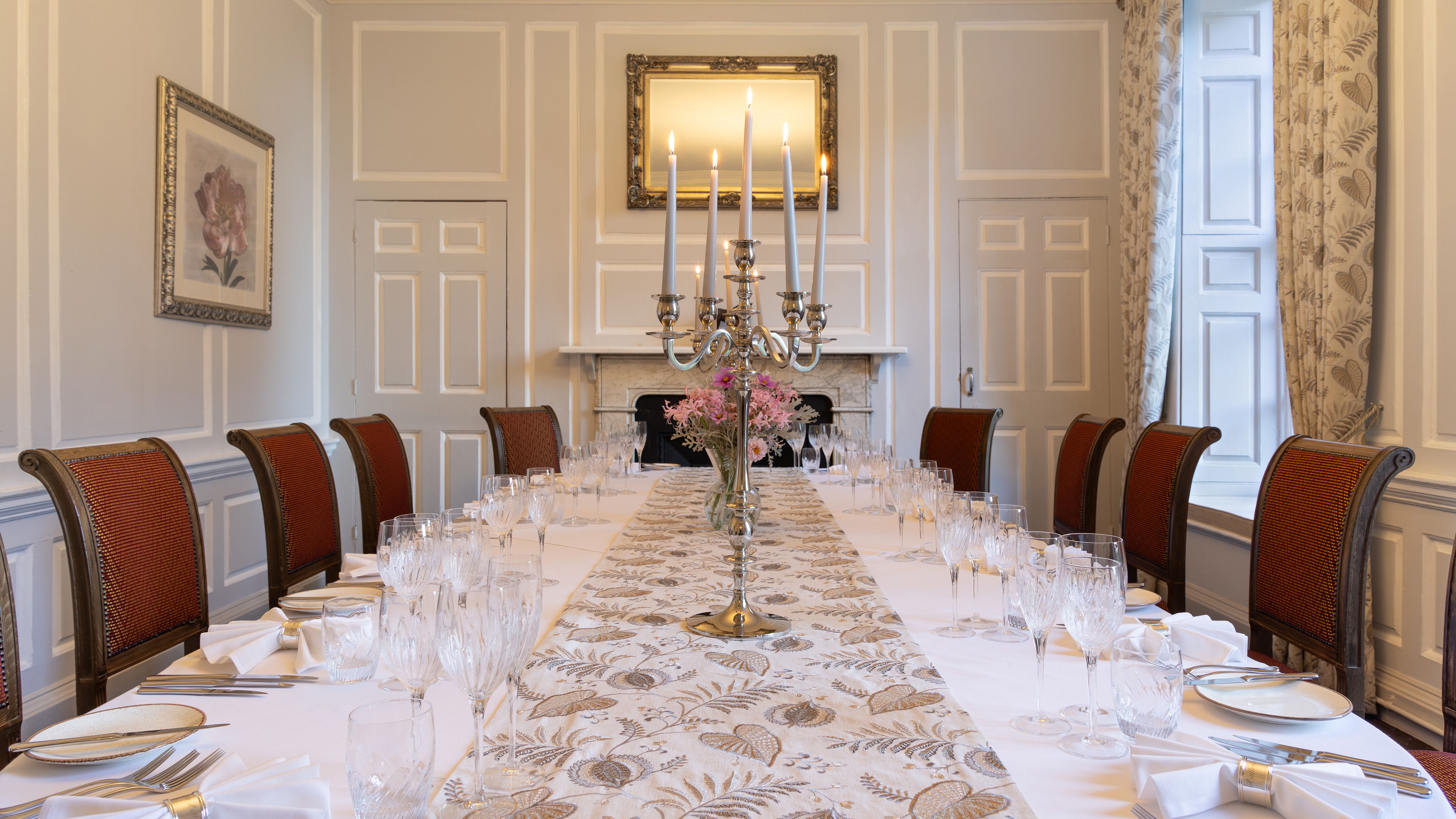 Elegant dining room with a long table set for a formal meal, featuring red chairs, white tablecloth, glassware, and a silver candelabra centerpiece.