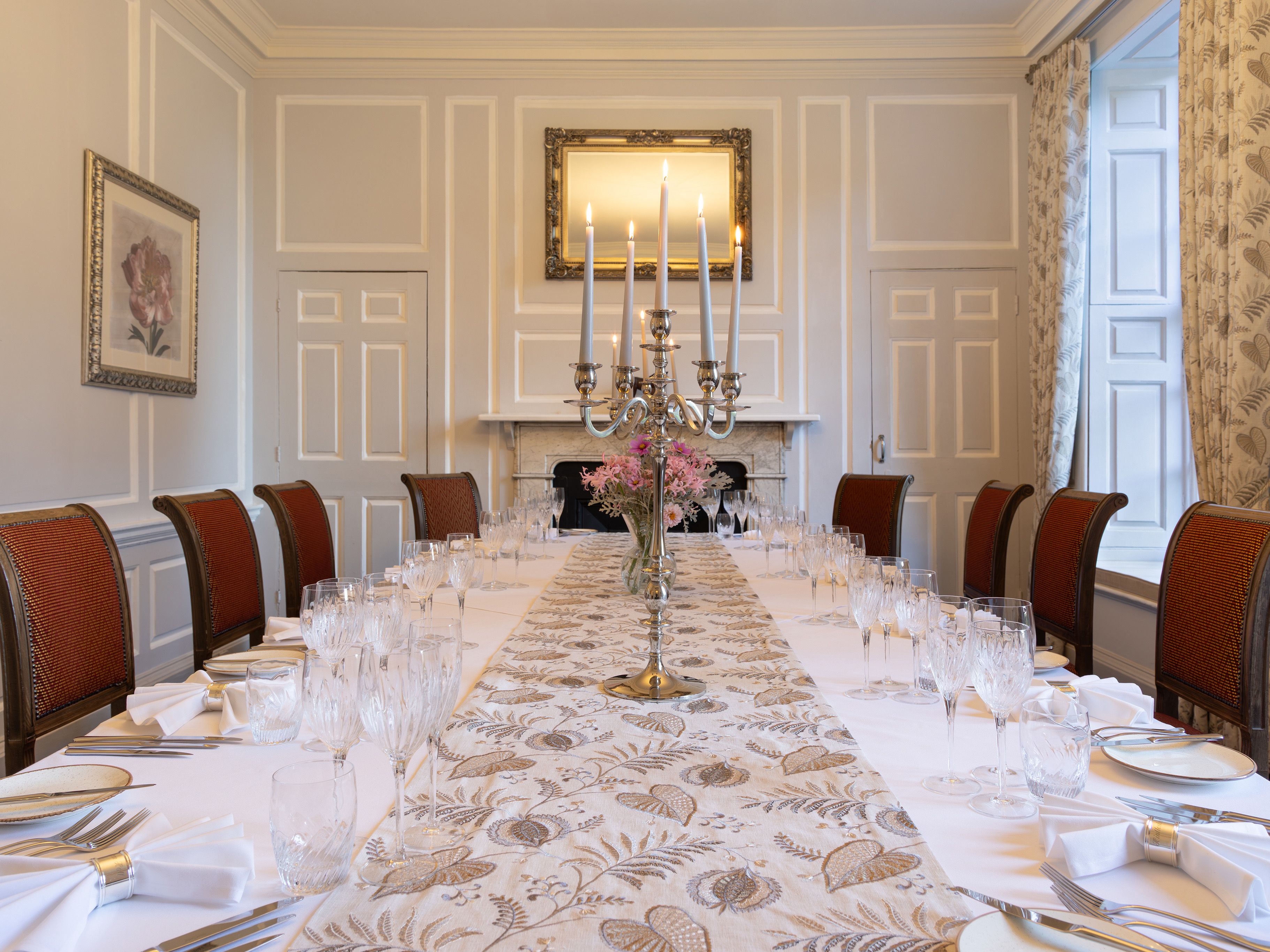 Elegant dining room with a long table set for a formal meal, featuring red chairs, white tablecloth, glassware, and a silver candelabra centerpiece.