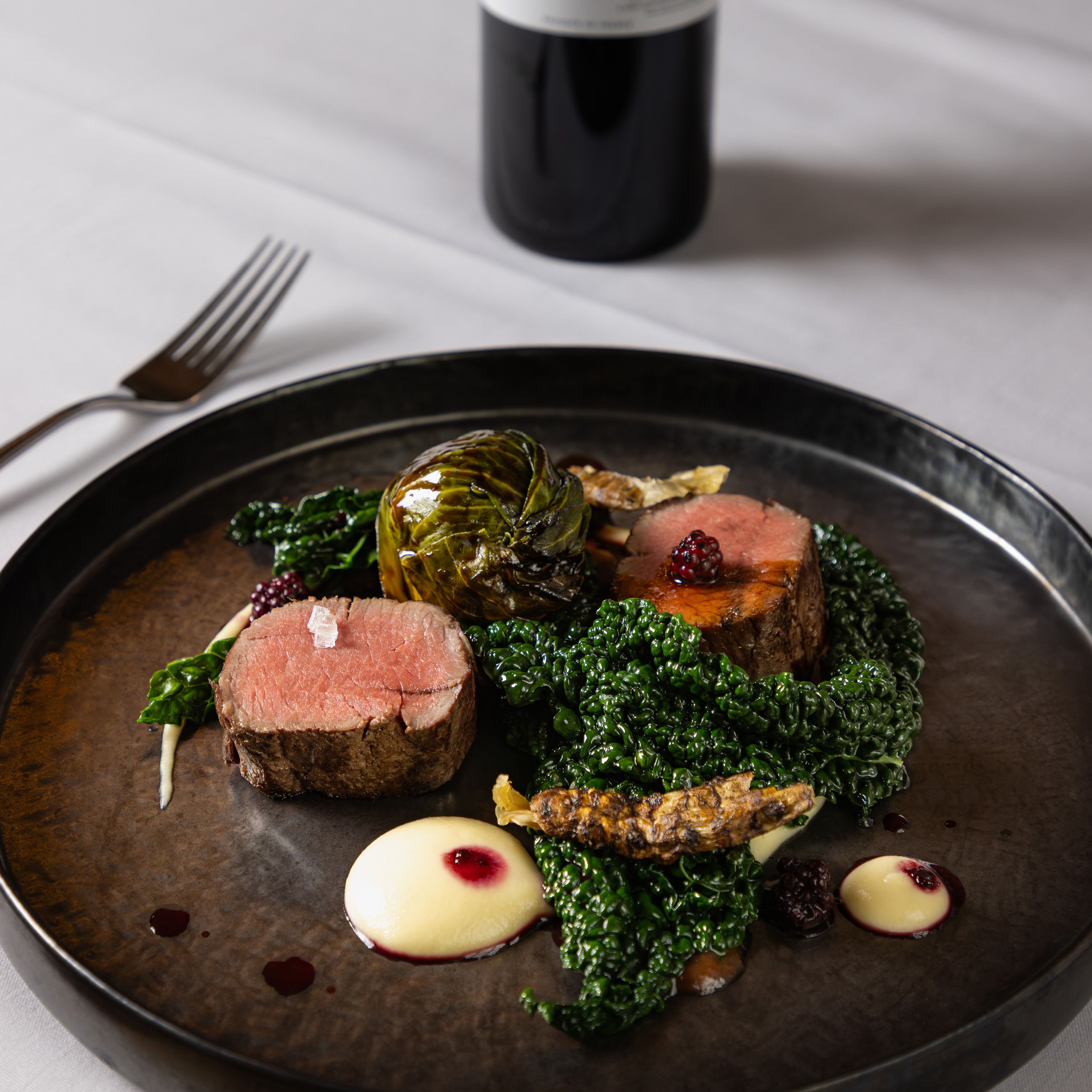 Gourmet plated beef dish with greens and sauce, served with a bottle of red wine