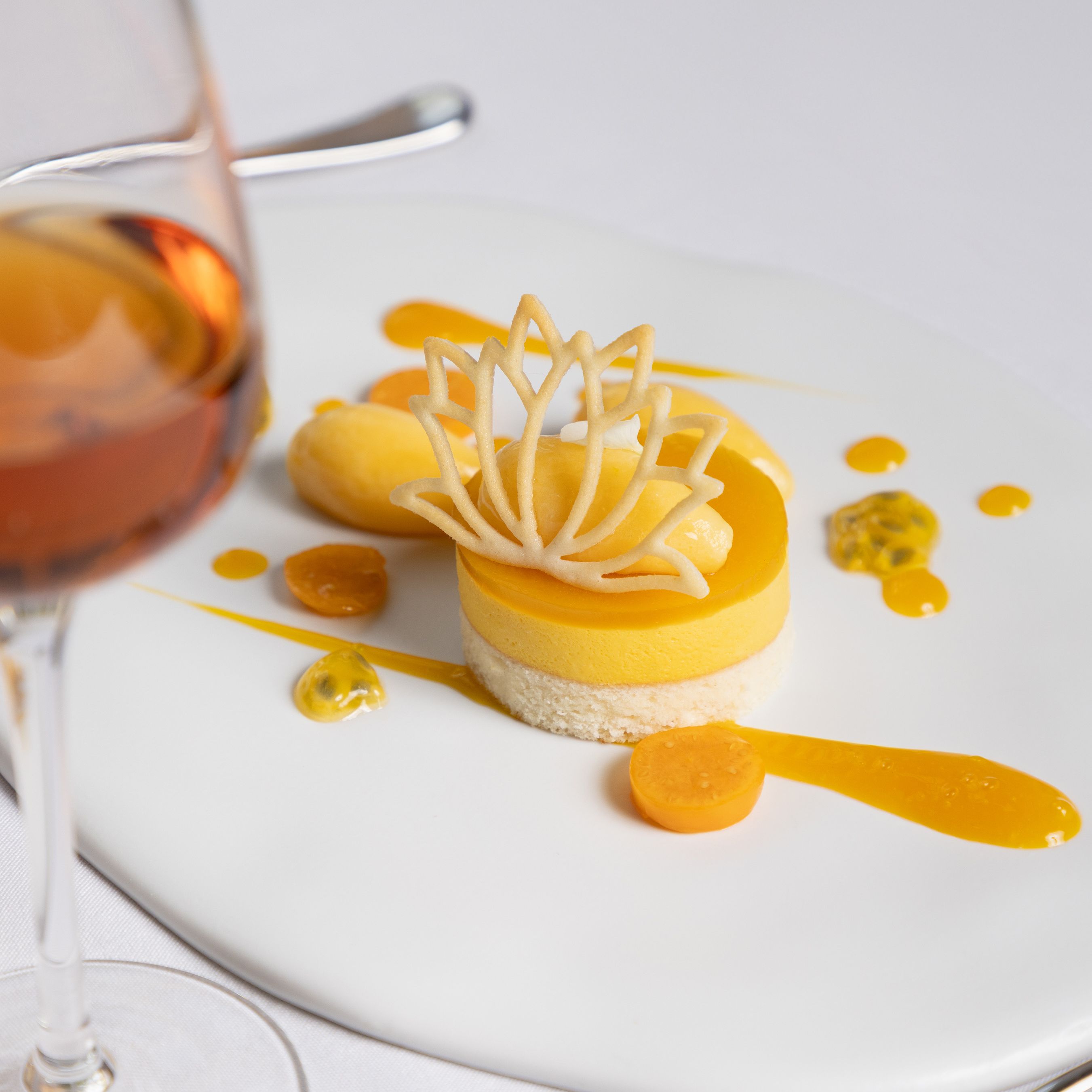 Elegant plated yellow dessert with decorative cookie art and fruit, accompanied by a glass of dessert wine.