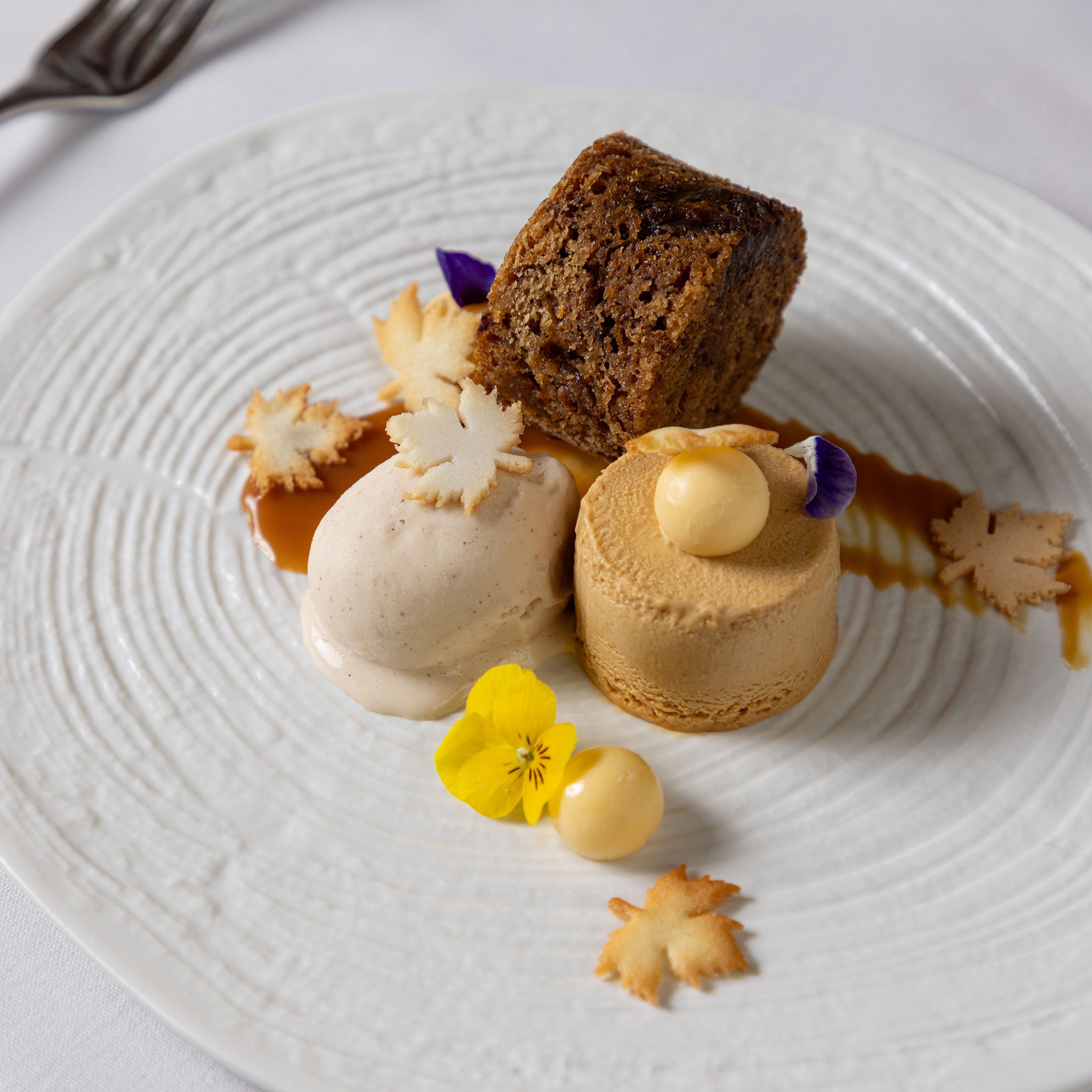 Elegant plated dessert with cake, ice cream, mousse, decorative leaves, and edible flowers