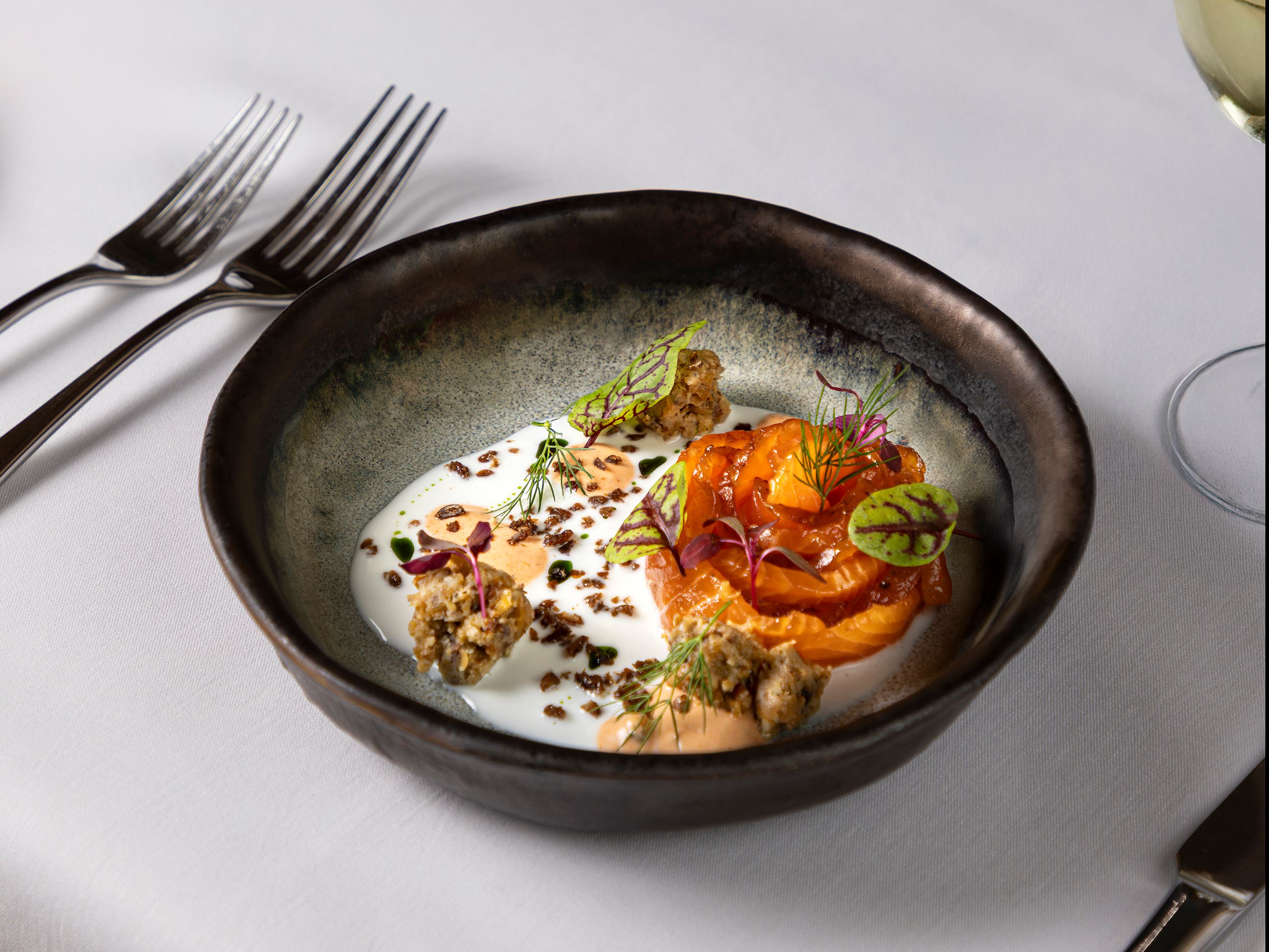 Gourmet plated dish of salmon with sauce, garnishes, and microgreens in a black bowl on a white tablecloth.