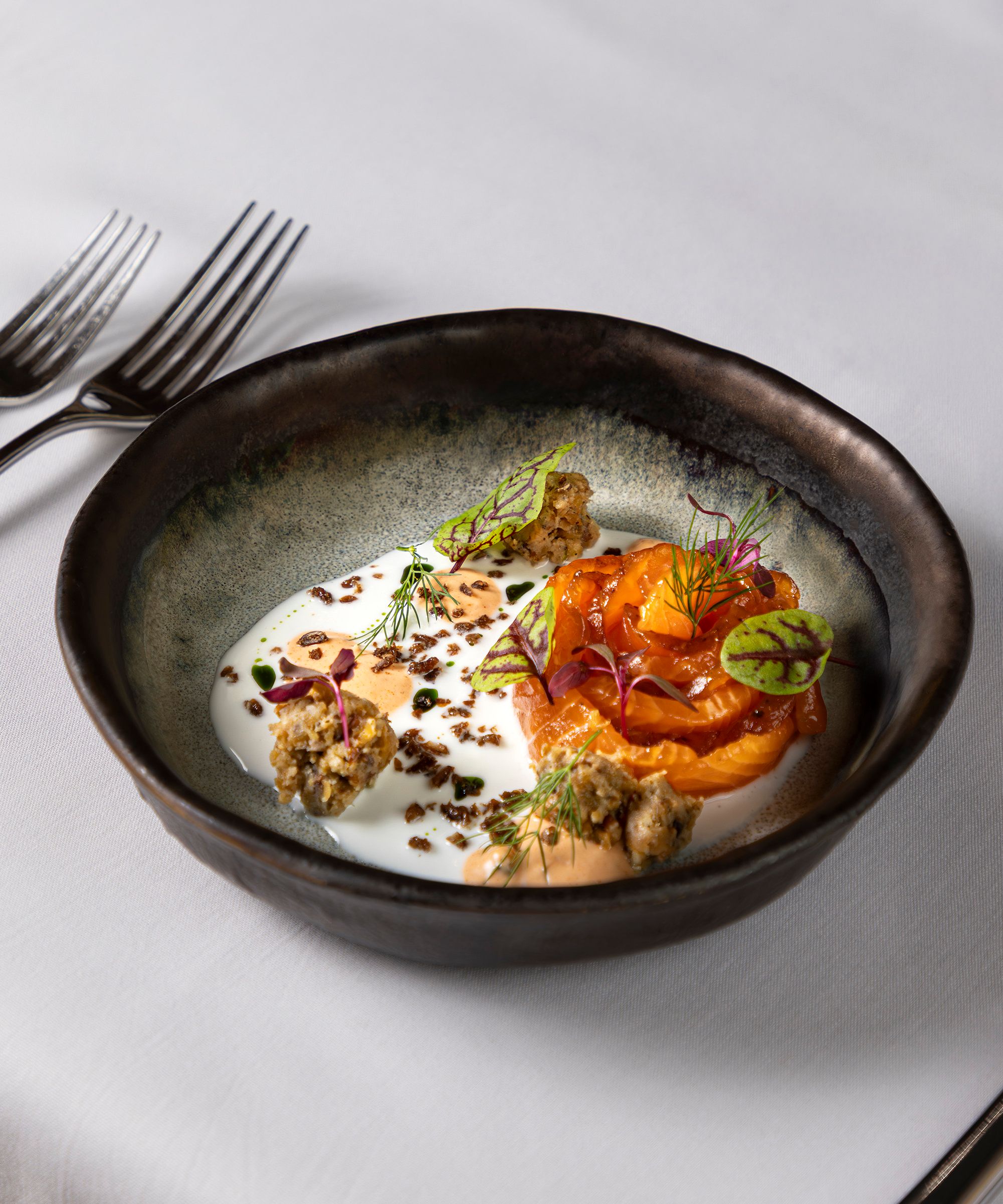 Gourmet plated dish of salmon with sauce, garnishes, and microgreens in a black bowl on a white tablecloth.