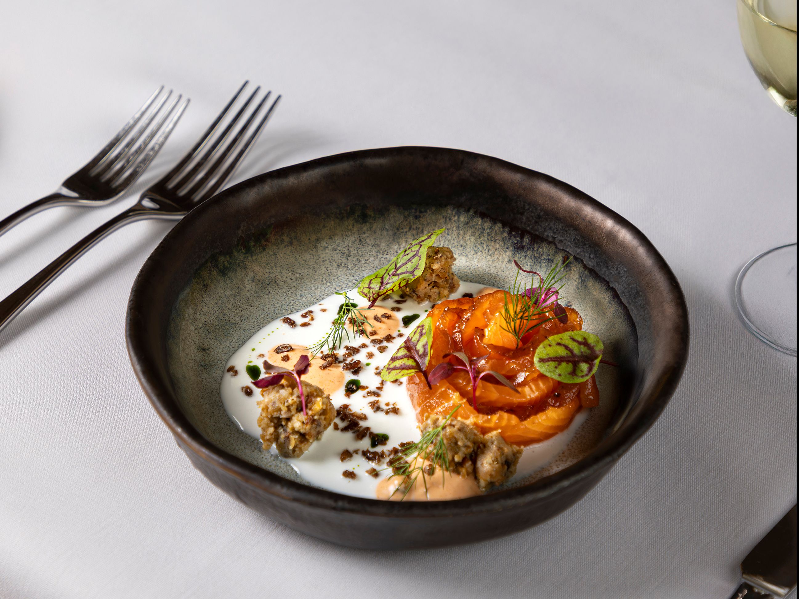 Gourmet plated dish of salmon with sauce, garnishes, and microgreens in a black bowl on a white tablecloth.