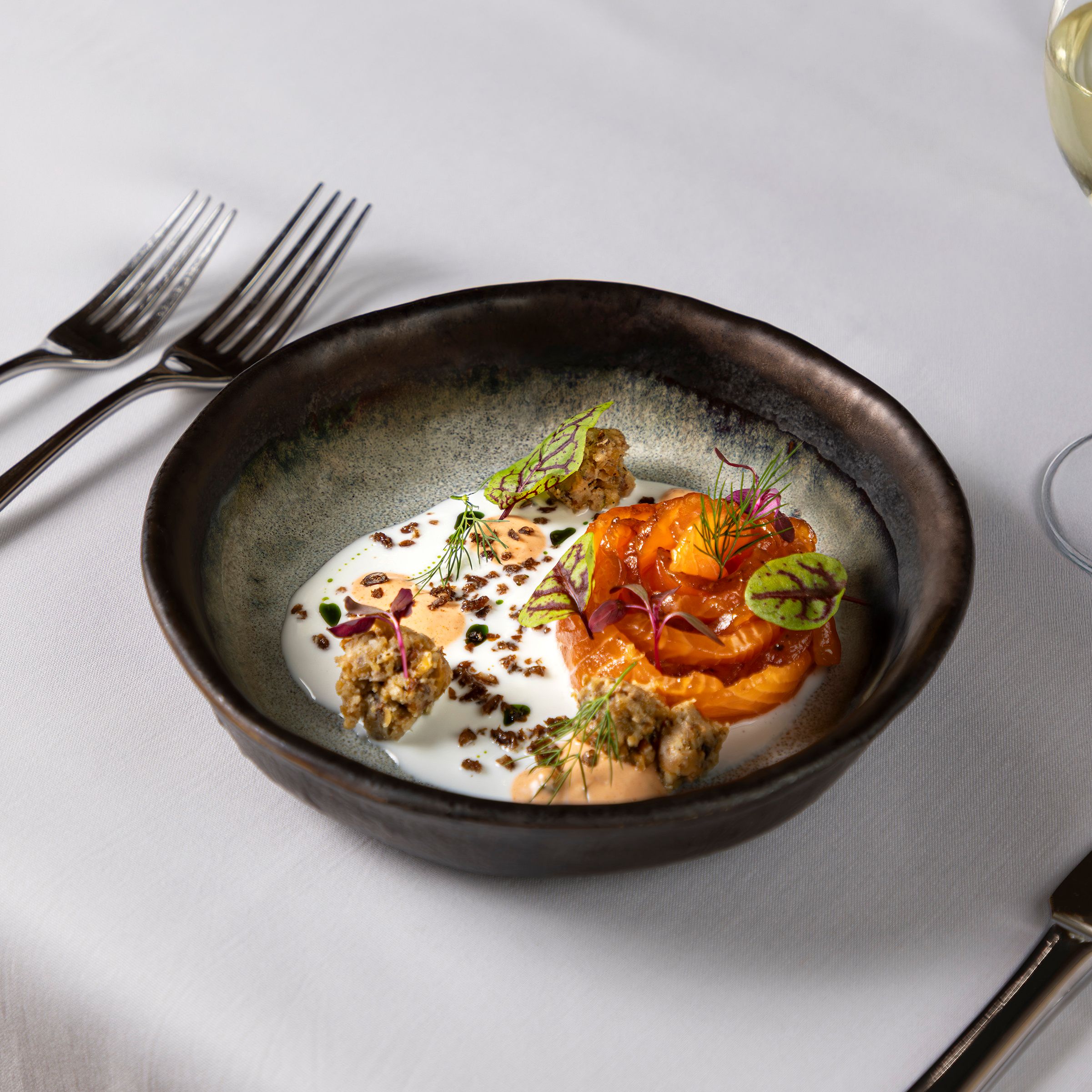 Gourmet plated dish of salmon with sauce, garnishes, and microgreens in a black bowl on a white tablecloth.
