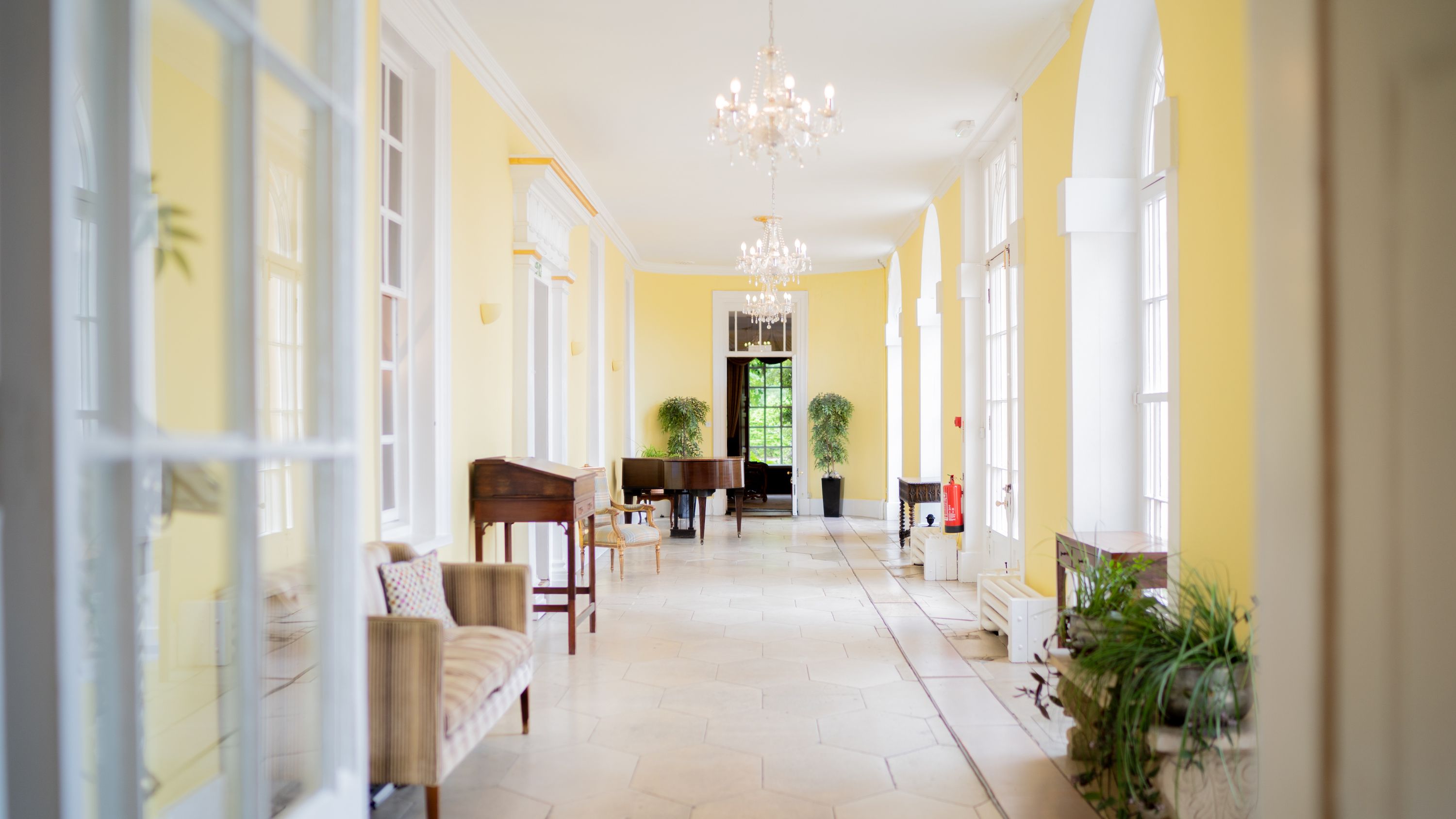 Bright, elegant hallway with yellow walls, white trim, chandeliers, large windows, and classic furniture.