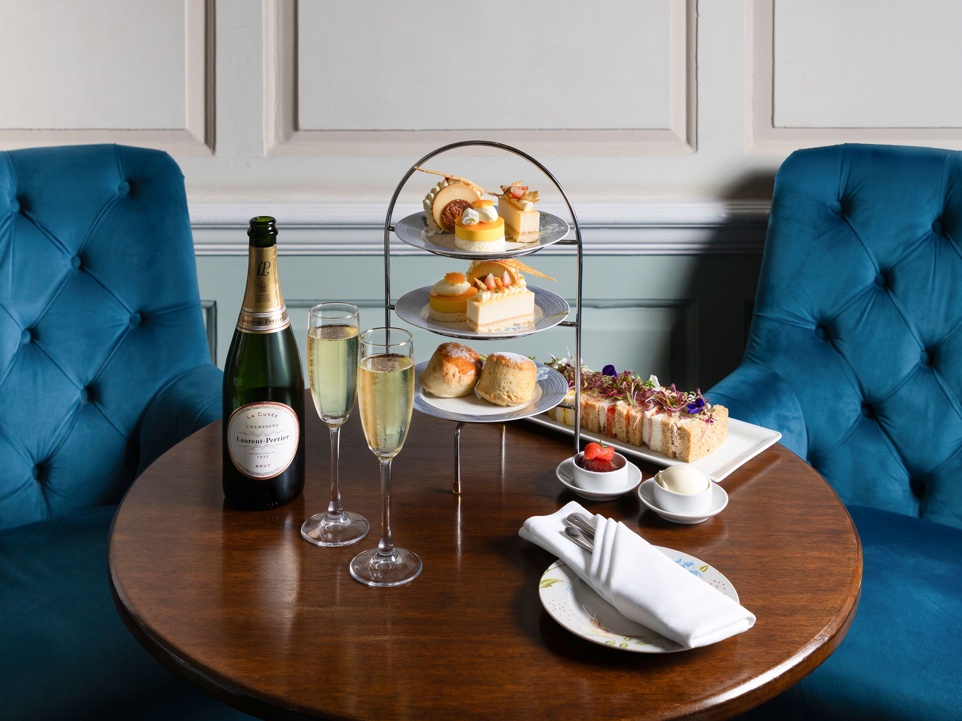 Afternoon tea set with pastries, scones, sandwiches, and champagne on a wooden table between two blue chairs