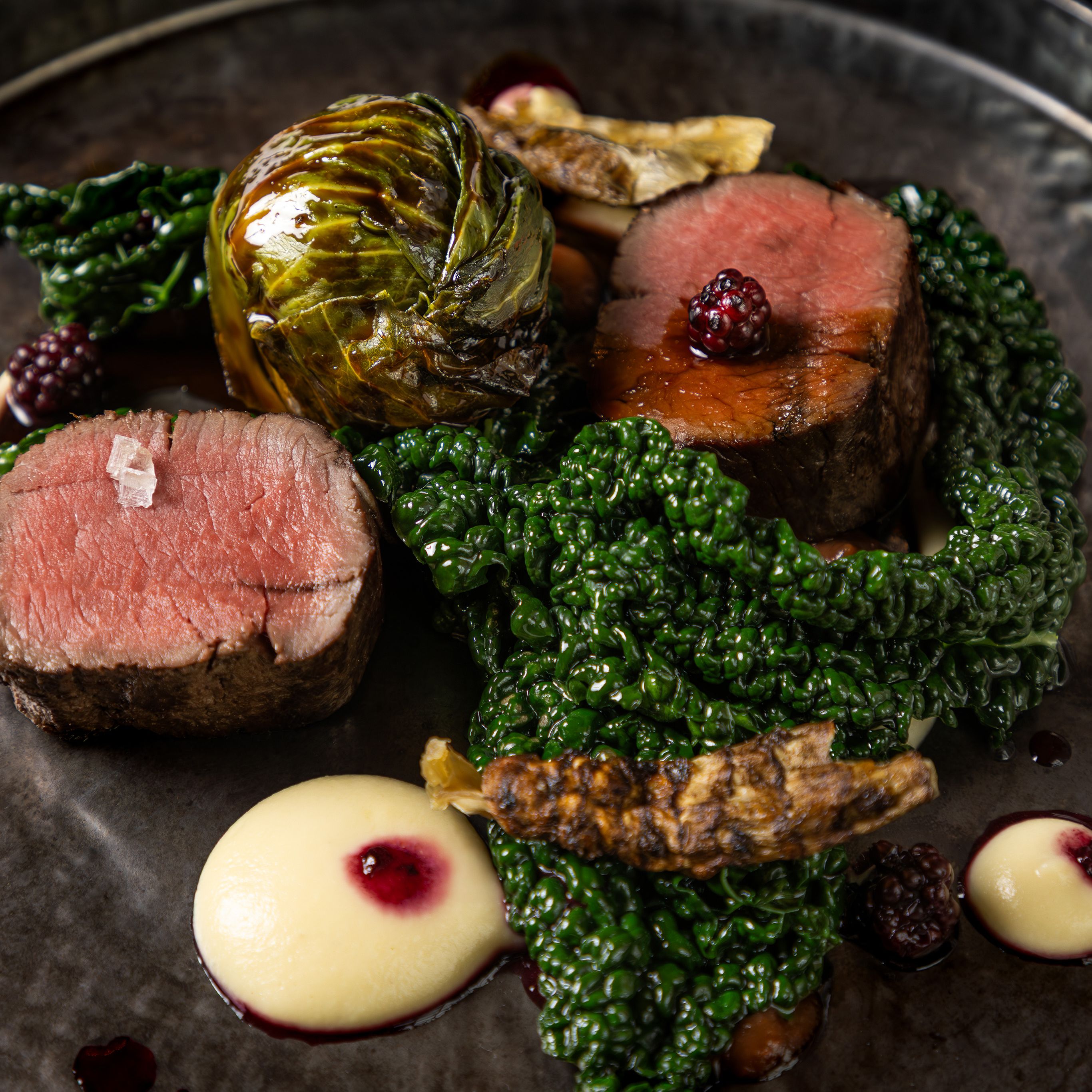 Gourmet plated dish with beef, leafy greens, creamy sauce, and blackberries
