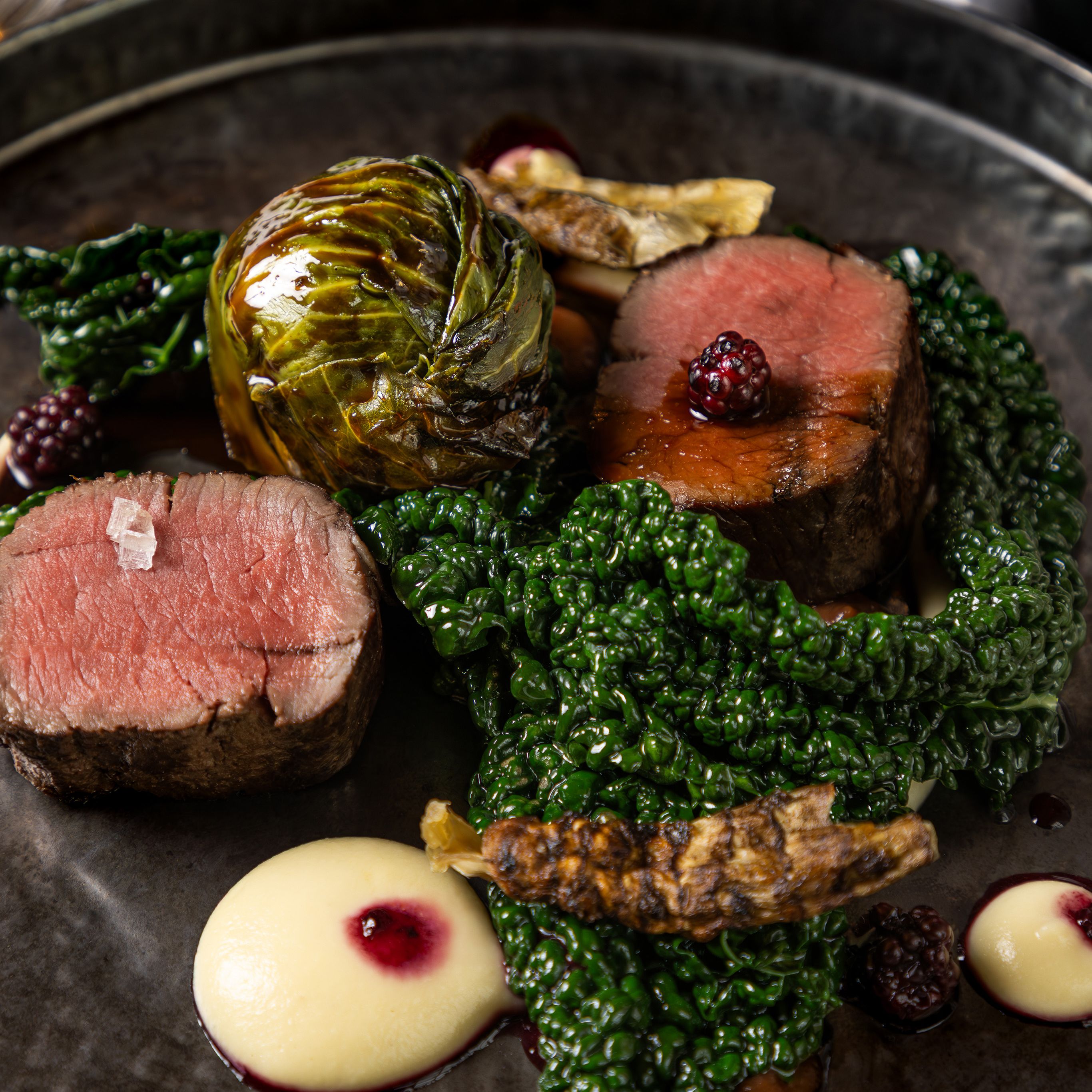 Gourmet plated dish with beef, leafy greens, creamy sauce, and blackberries