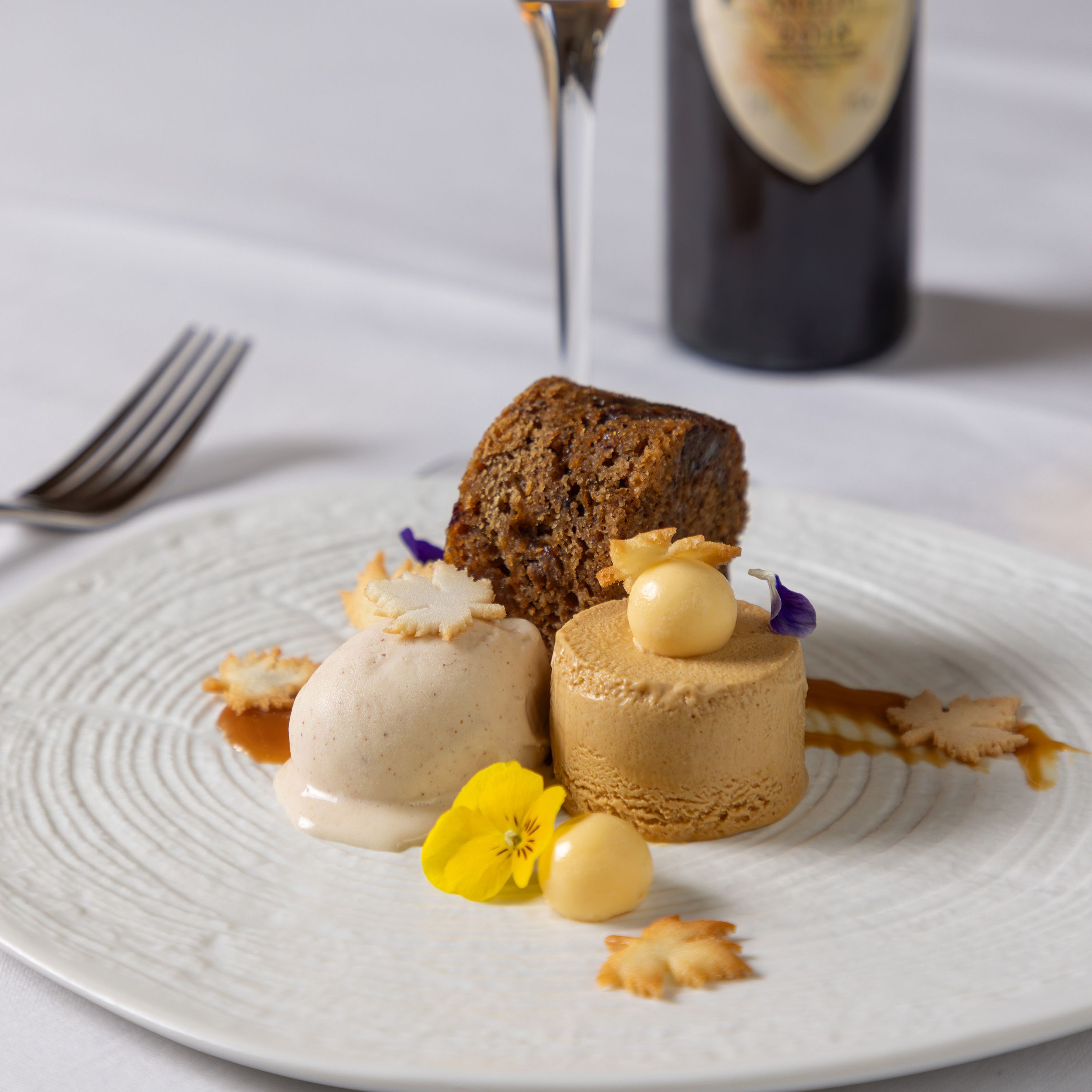 A plated dessert with cake, ice cream, and mousse, garnished with edible flowers and cookie pieces, next to a glass of dessert wine and a wine bottle.