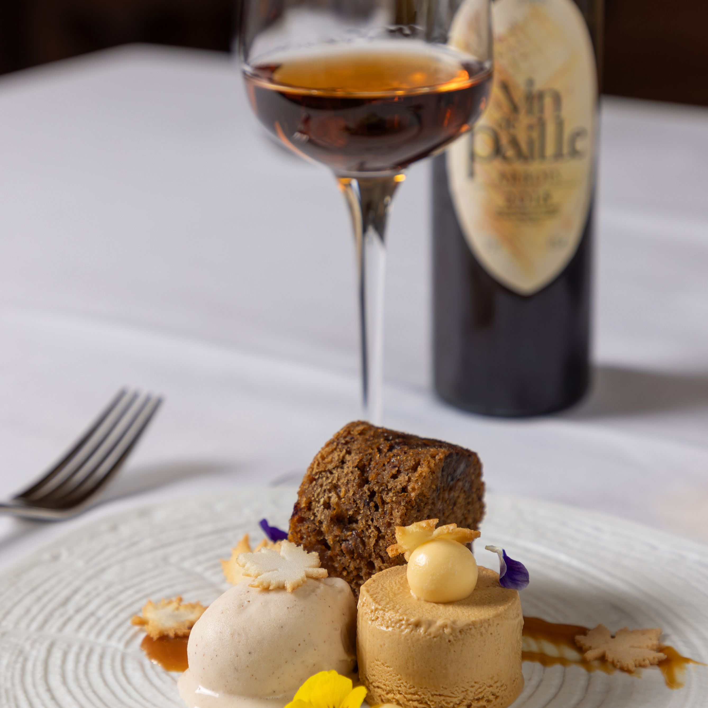 A plated dessert with cake, ice cream, and mousse, garnished with edible flowers and cookie pieces, next to a glass of dessert wine and a wine bottle.