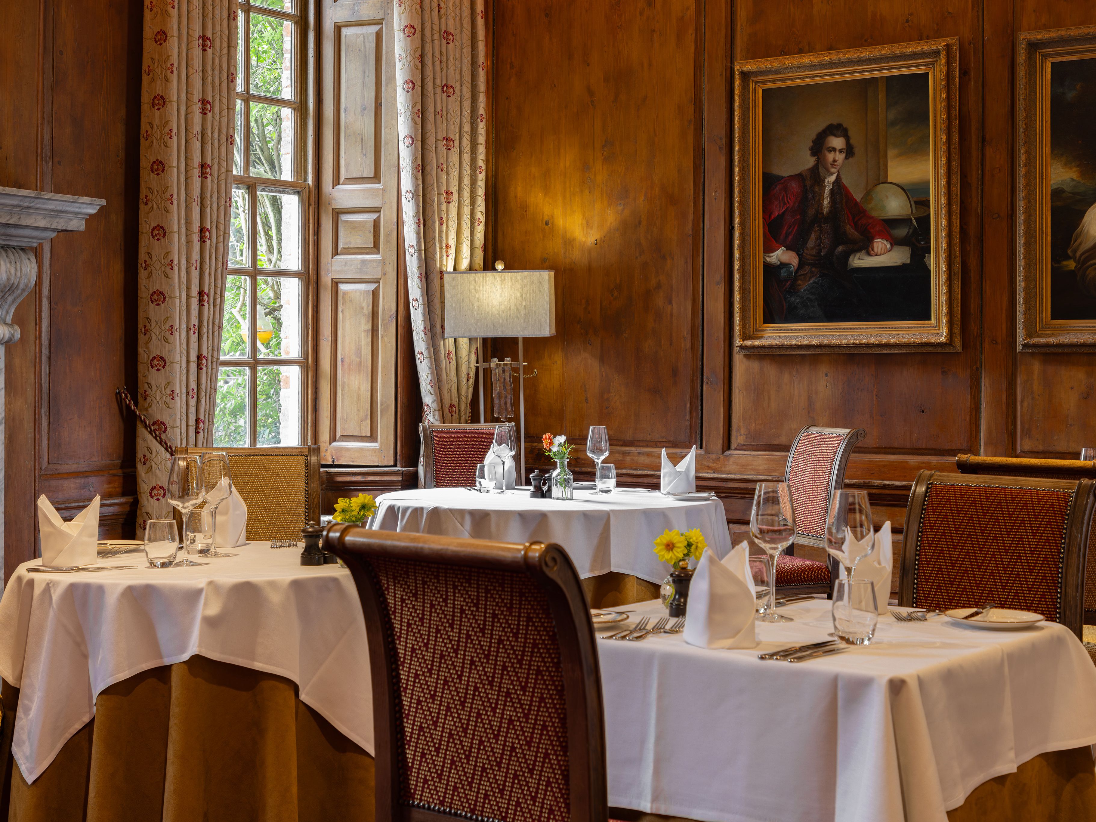 Elegant restaurant dining room with white tablecloths, ornate chairs, and classic paintings on wooden-paneled walls.